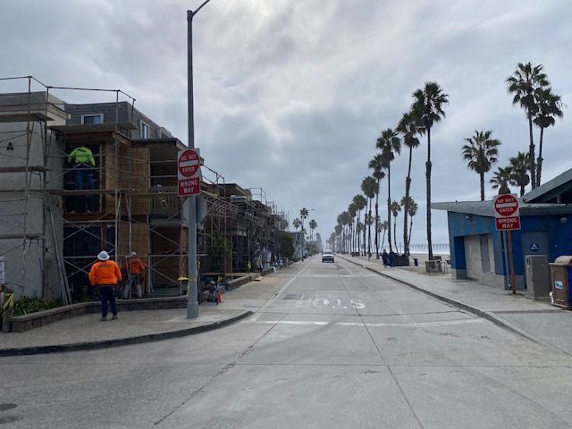 Palm-lined street with construction scaffolding on the left and blue buildings on the right under a cloudy sky