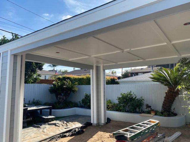 Covered patio with white columns and ceiling fans in a landscaped backyard, with a ladder on the ground.