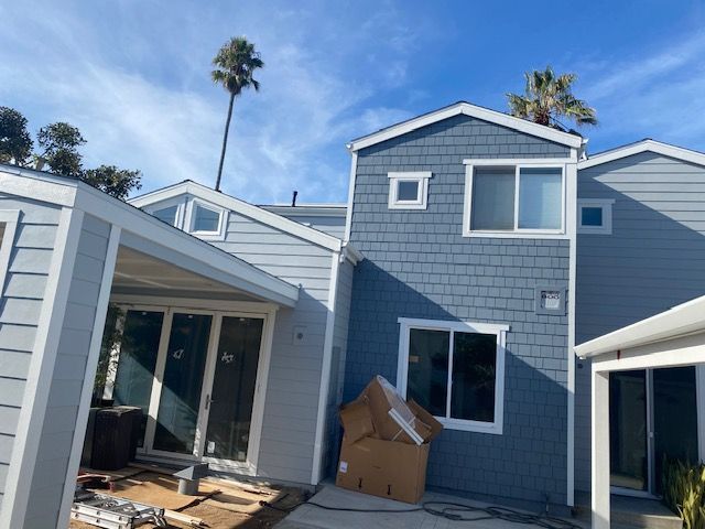 Blue-sided house under construction with open sliding doors, cardboard boxes, and palm trees against a sunny sky