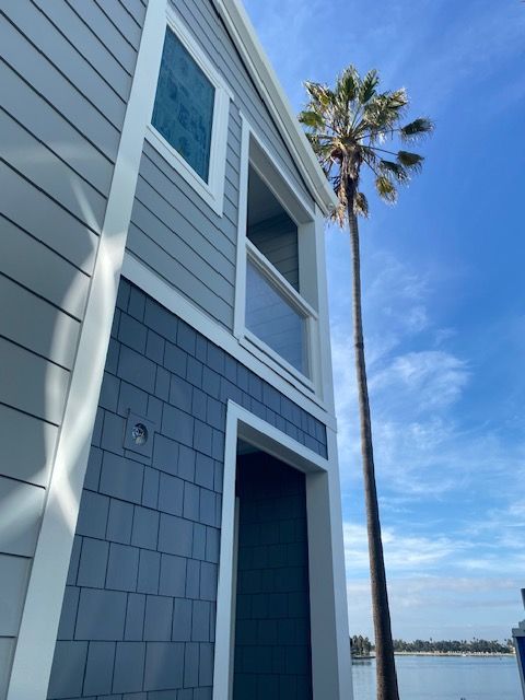 Blue-gray coastal house beside water under a clear sky, with a tall palm tree in the background