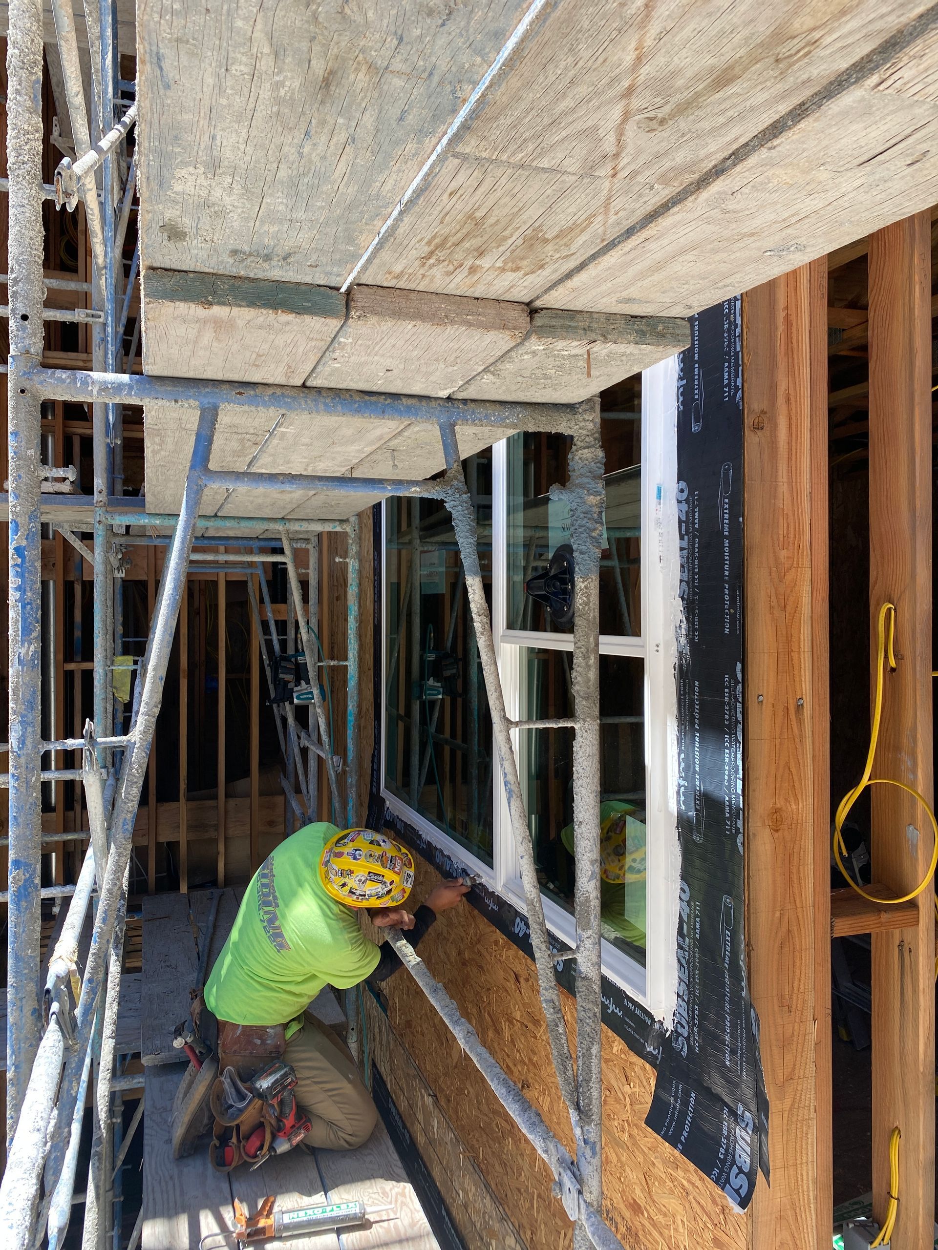 Construction worker kneeling on scaffolding, installing window trim inside a framed building under construction.