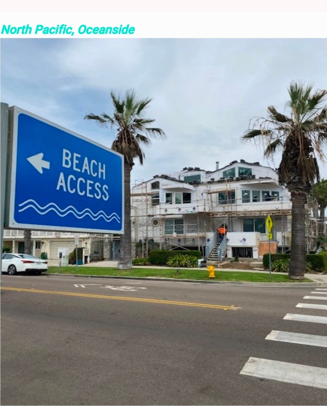Blue beach access sign on a street corner near palm trees and a white building in Oceanside.