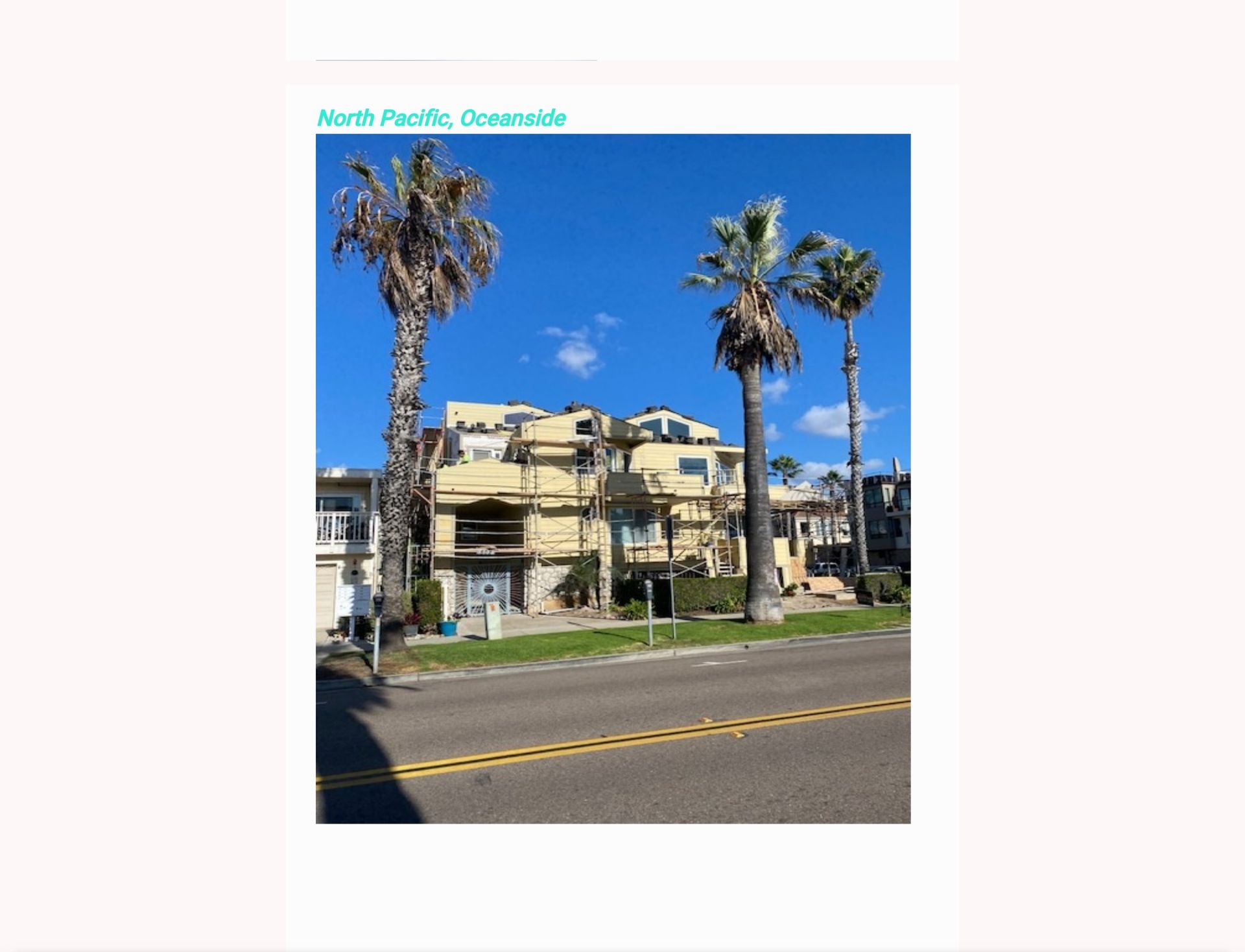 Palm trees frame a beige apartment building under a clear blue sky beside a road.