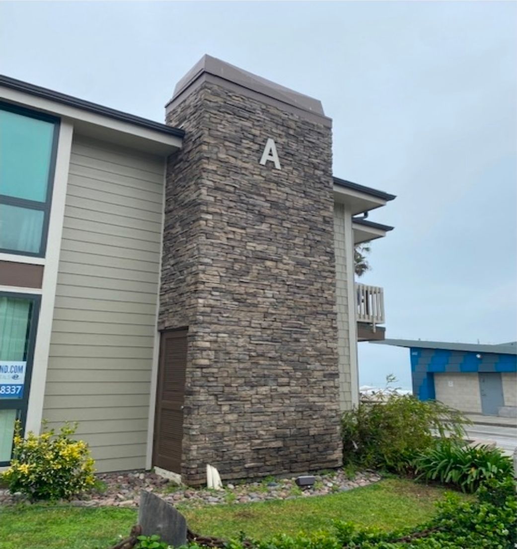 Stone apartment building entrance with large letter A and landscaped shrubs under a cloudy sky