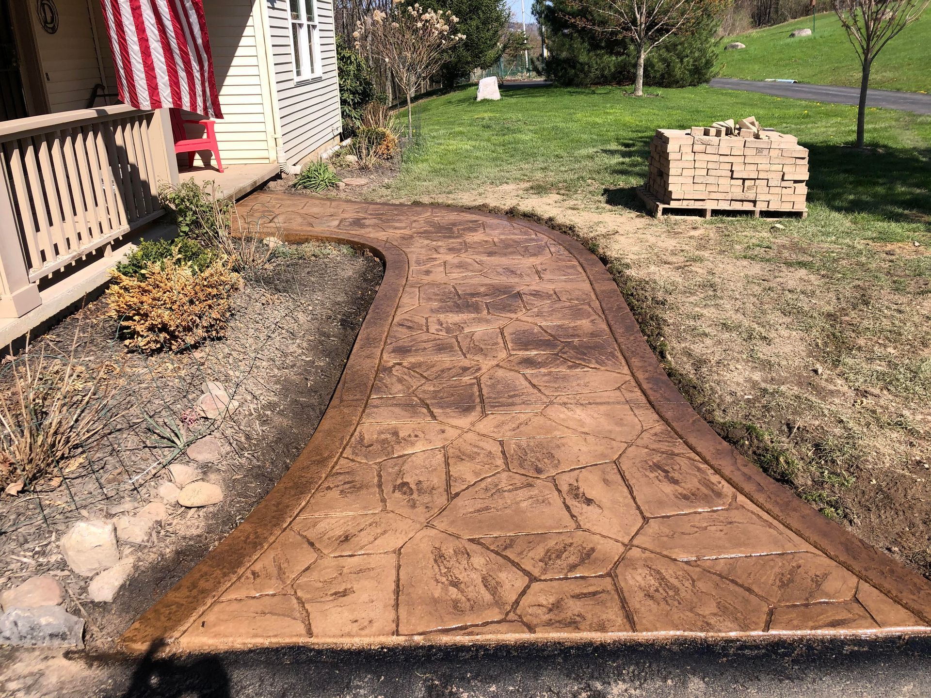 Brown stamped concrete walkway curving from a house with a US flag to the street.