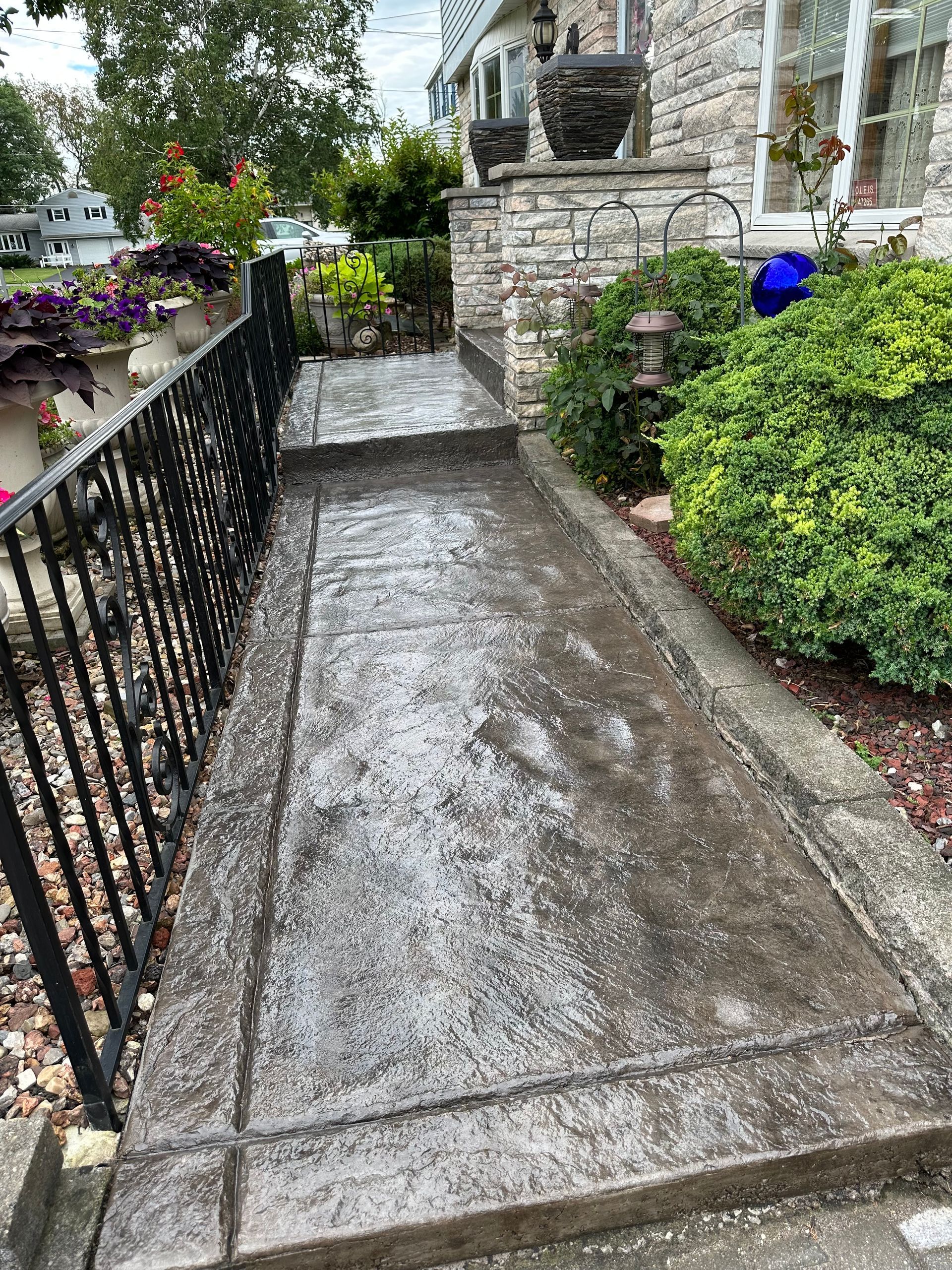 Concrete walkway with a decorative border, leading to a stone house entrance with bushes and a railing.