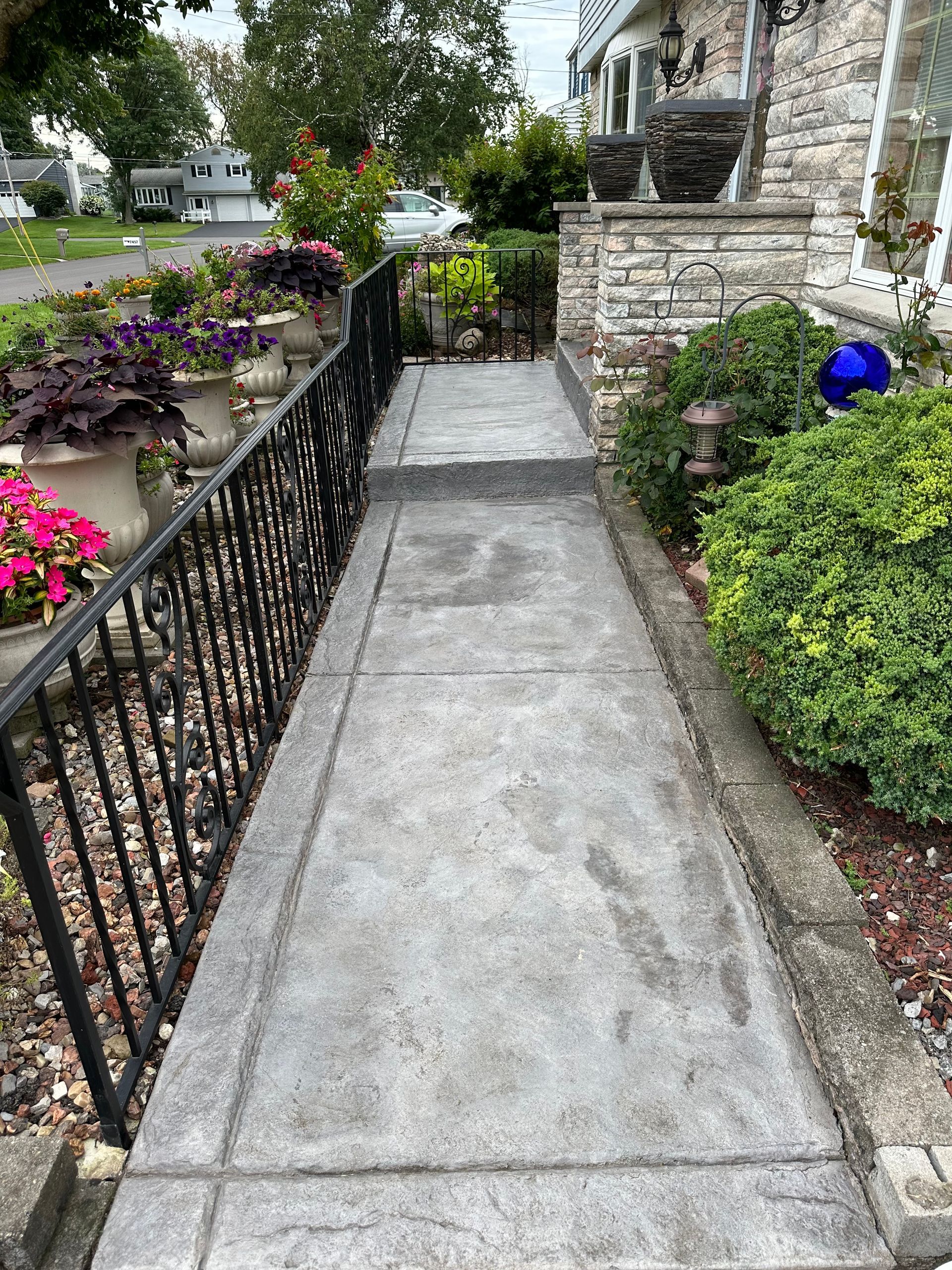 Concrete walkway lined with black railing and flowerbeds leading to a stone-covered house entrance.