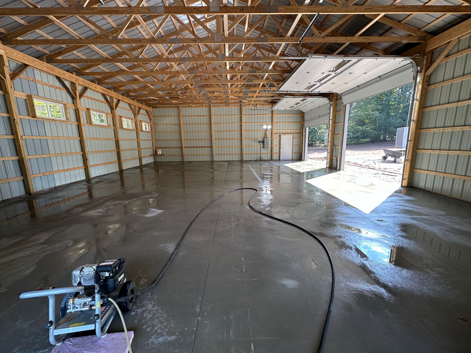 Newly poured concrete floor in a large open building with wooden beams and a partially open garage door.