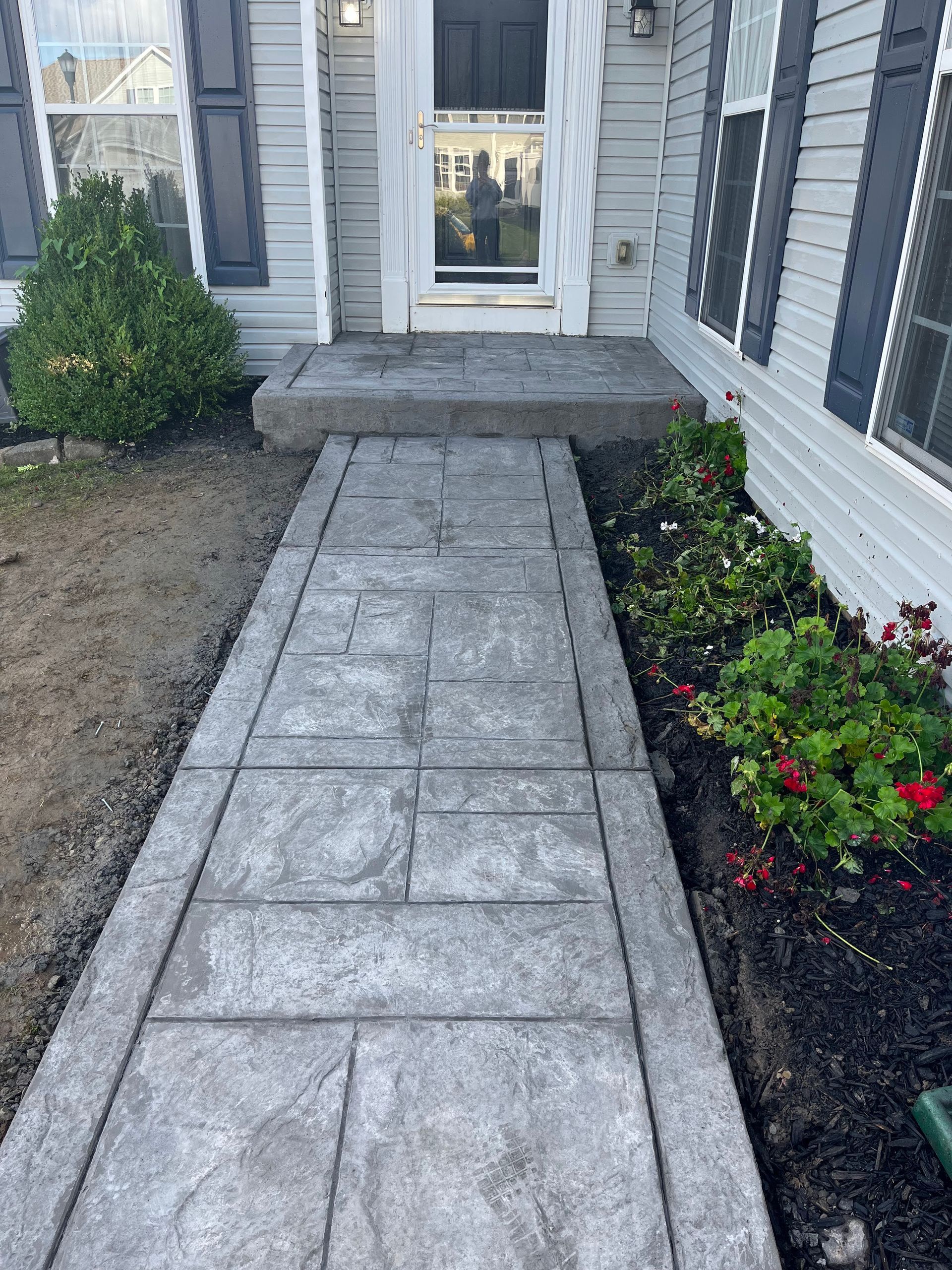 Concrete walkway leading to a house entrance with steps. Grey concrete, dark mulch, and green plants.