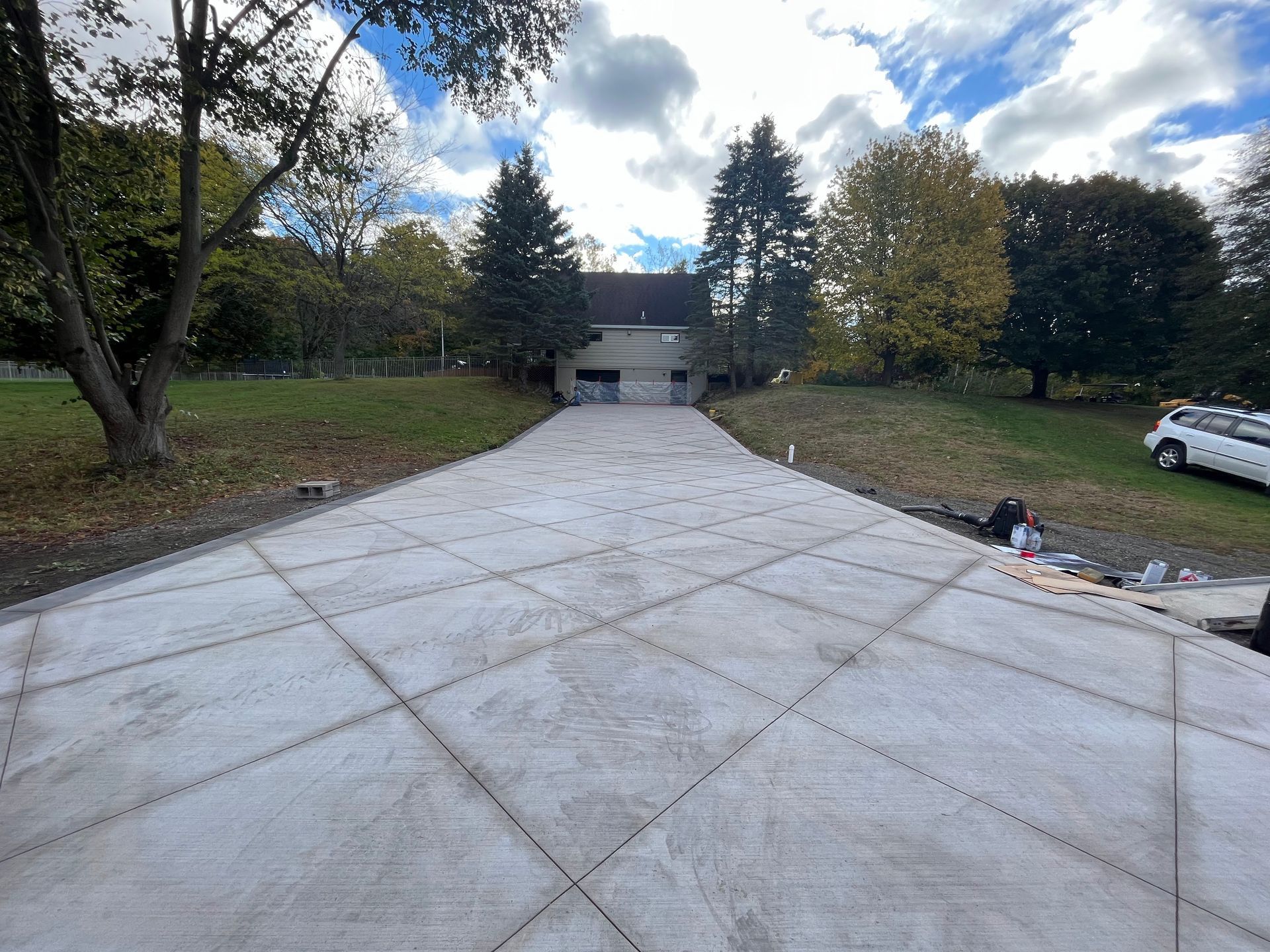 A wide concrete driveway leading to a house, surrounded by trees under a cloudy sky.