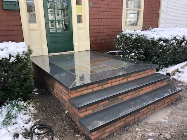 Brick steps and porch with dark stone surface, leading to a green door, snow on sides.