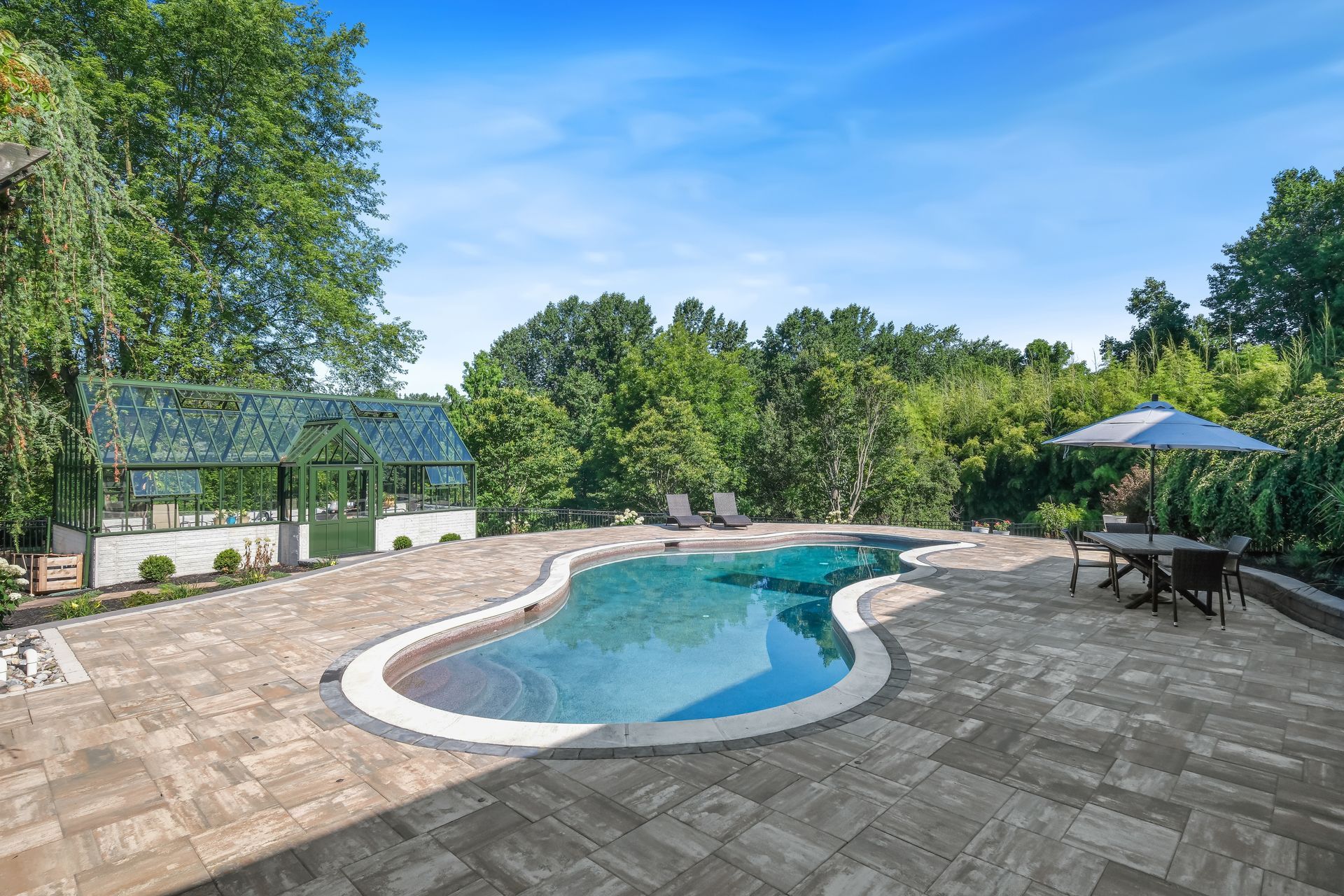 Backyard swimming pool with stone patio, umbrella table, and lush green trees under a blue sky