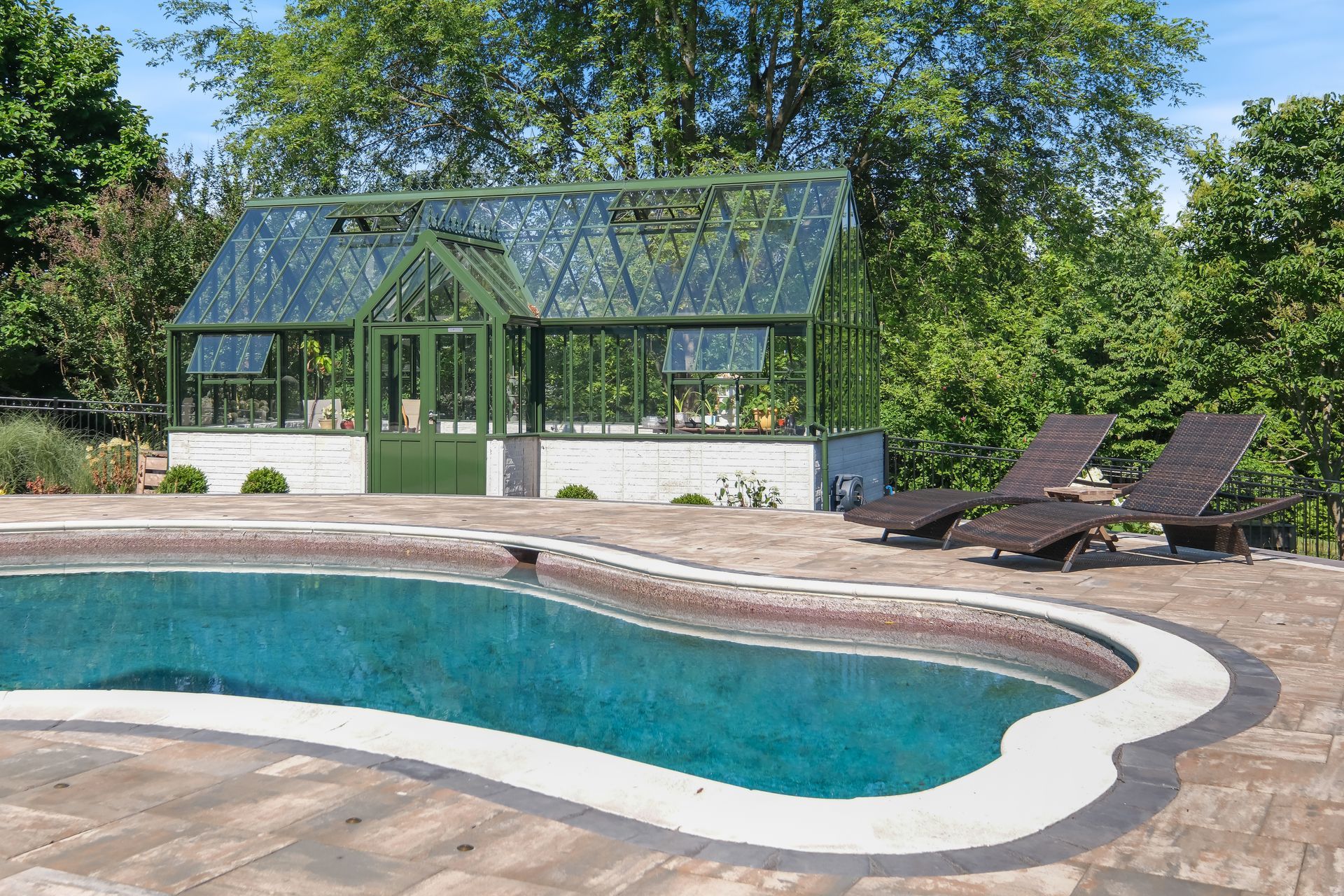 Curved backyard pool beside a glass greenhouse and lounge chairs under trees