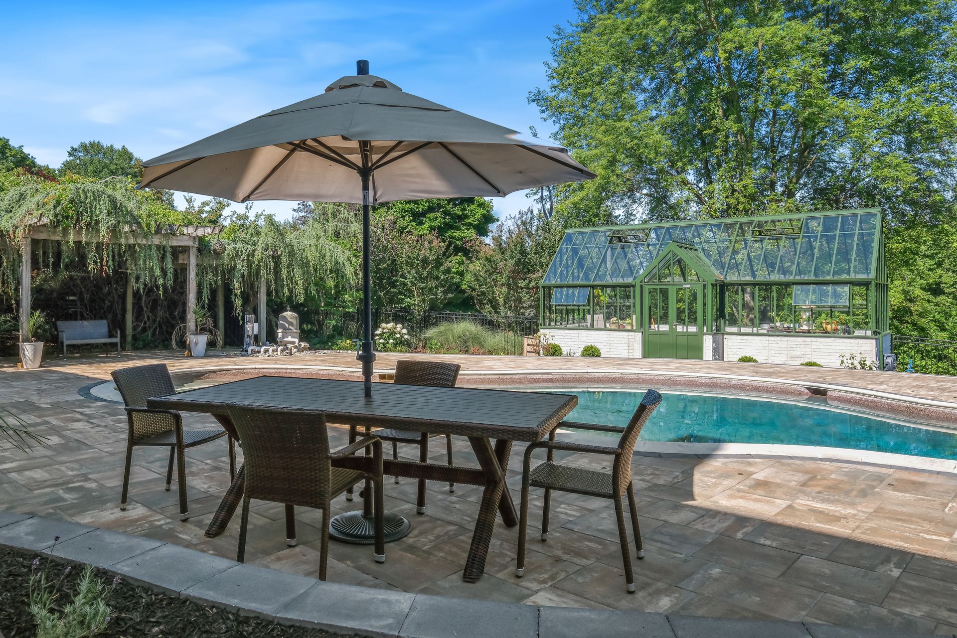 Patio with table, chairs, and umbrella beside a swimming pool in a sunny garden