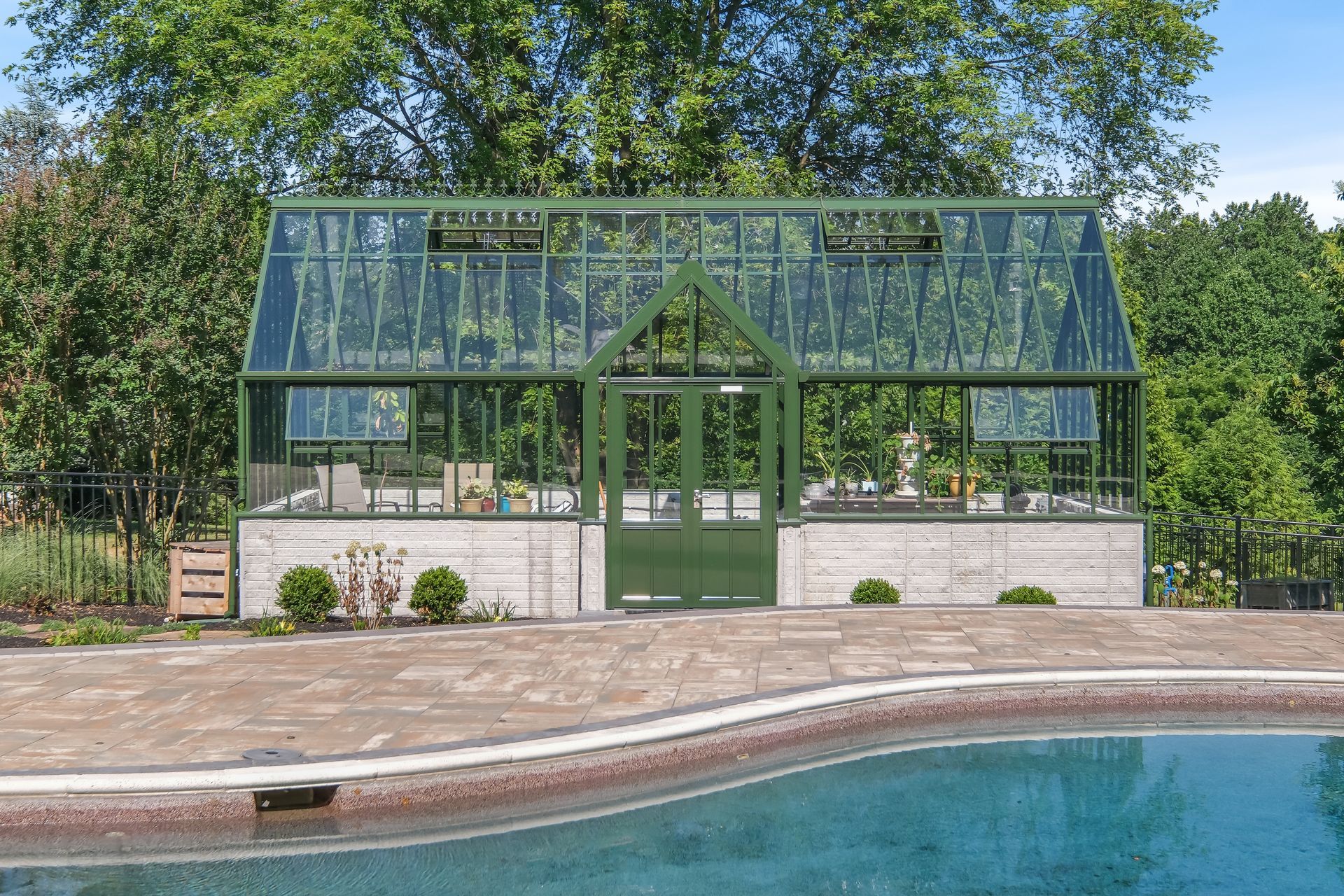 Green glass greenhouse by a pool, surrounded by trees and a stone patio