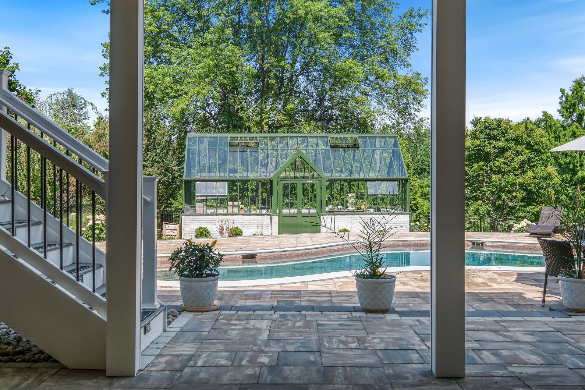 Covered patio overlooking pool and glass greenhouse, with stone flooring and blue sky beyond trees