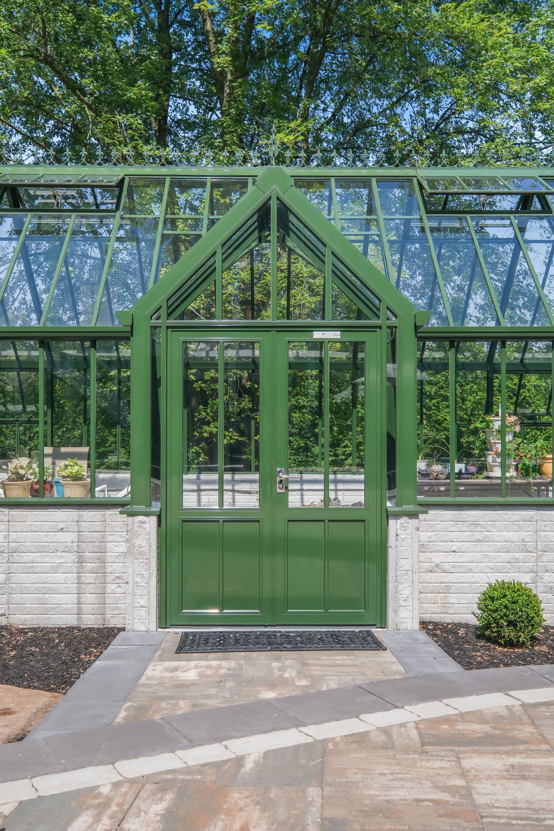 Green glass greenhouse entrance with double doors and metal frame, surrounded by trees and brick walls