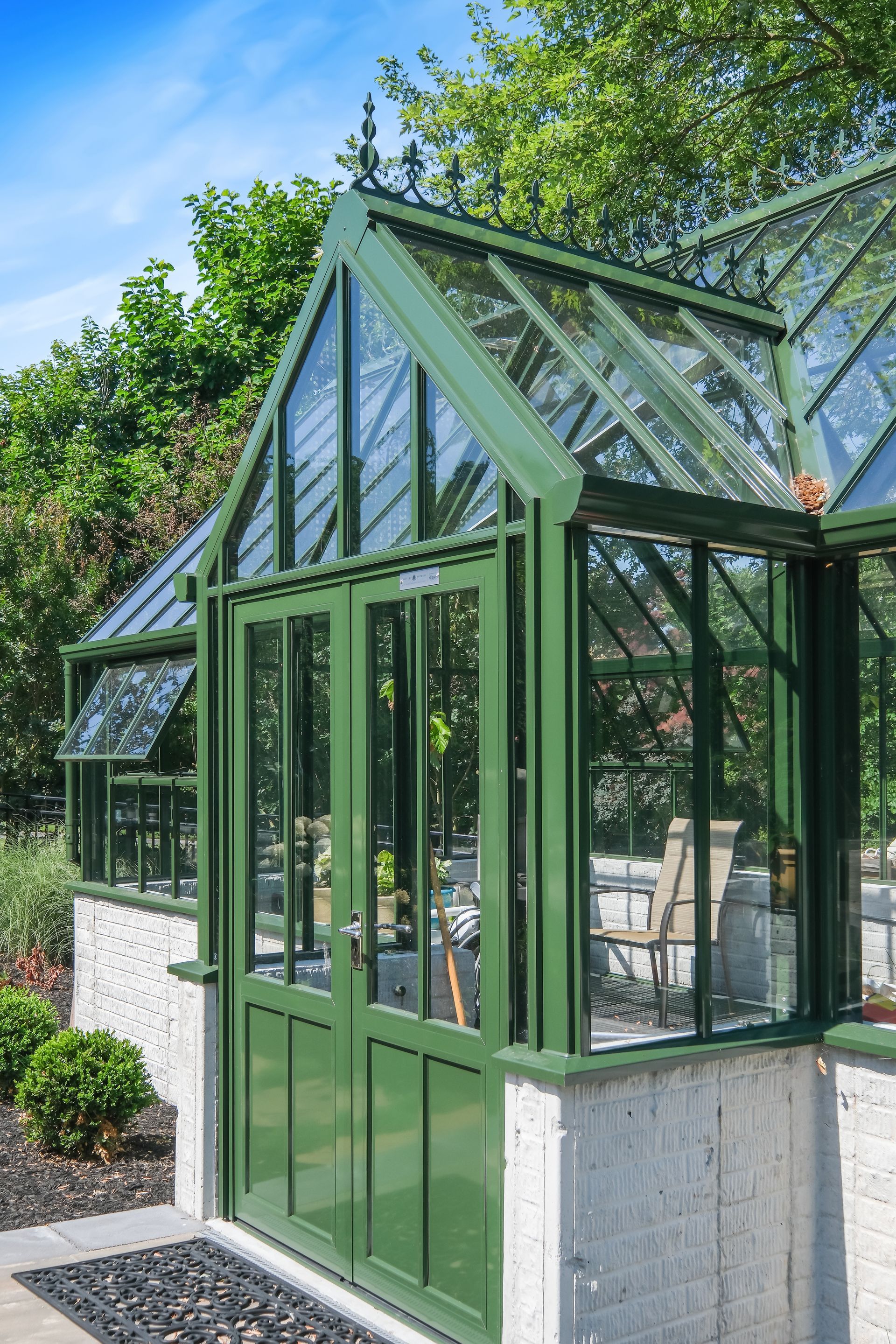 Green glass conservatory with black metal framing and white stone base in a leafy garden
