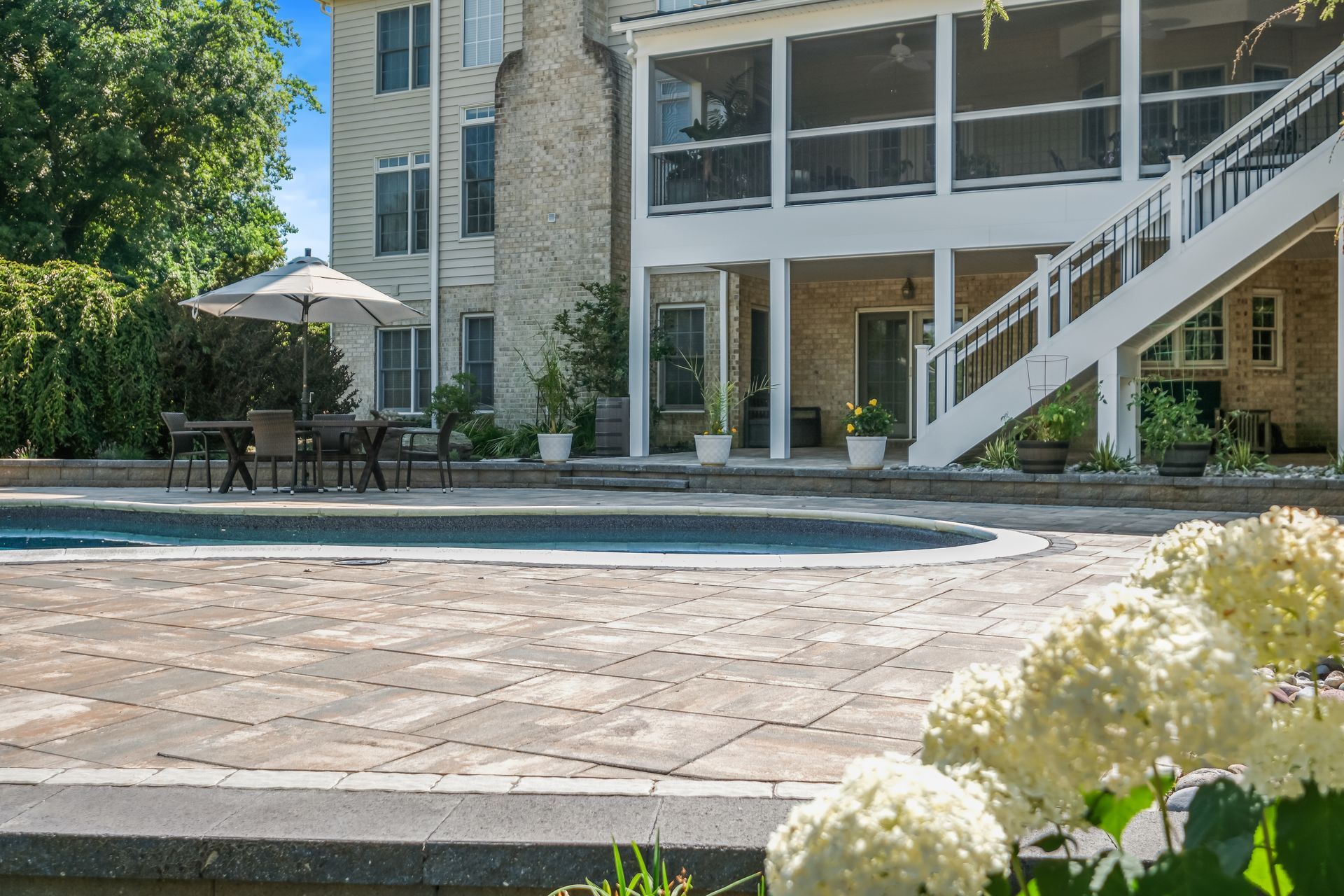 Patio with pool in front of a stone house, patio table and umbrella, and white flowers in the foreground