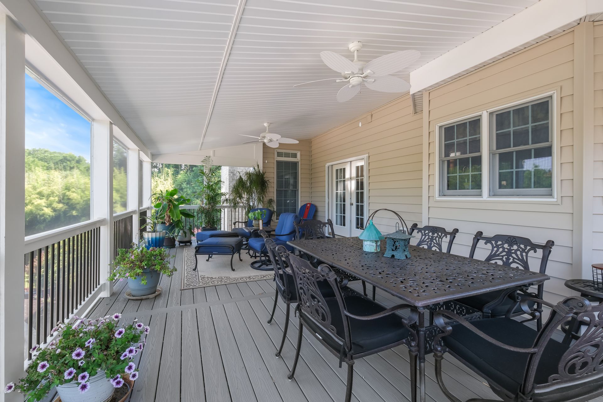 Covered porch with outdoor dining table, chairs, potted plants, and railing overlooking greenery
