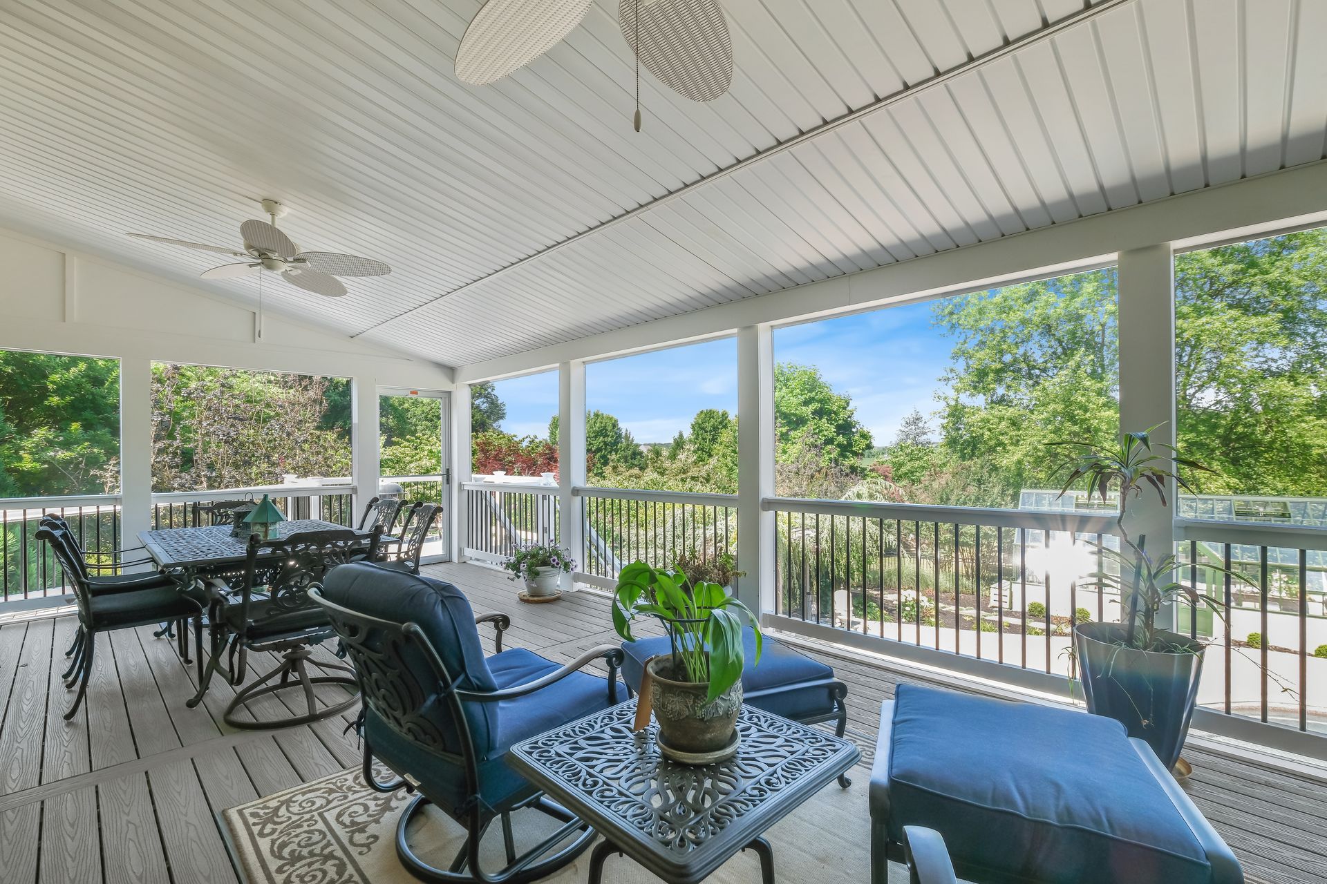 Bright screened porch with wicker seating, blue cushions, potted plants, and a view of trees outside