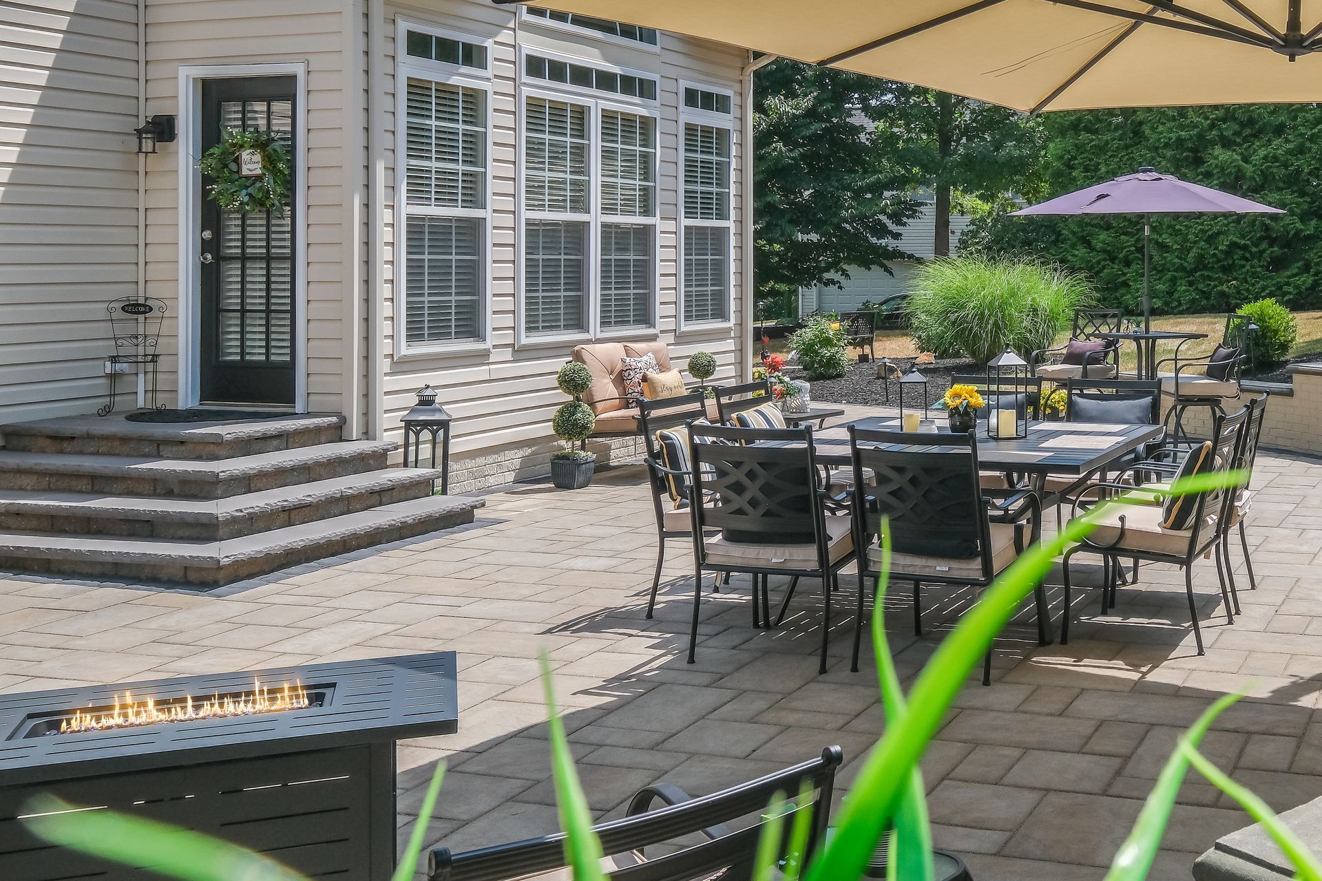 Patio with black metal tables and chairs beside a stone house, under large beige umbrellas.
