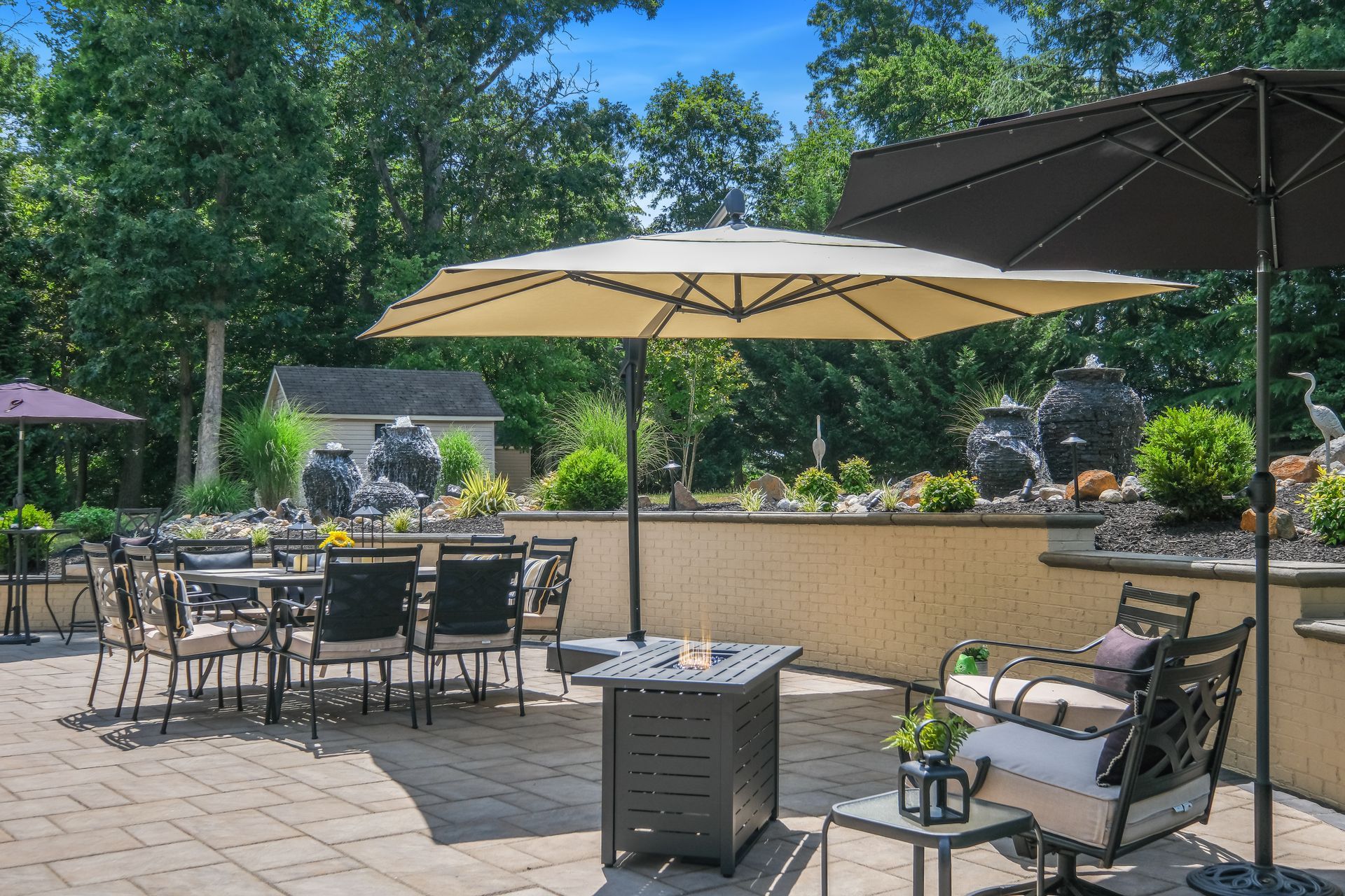 Outdoor patio with tables, chairs, umbrellas, and a fountain in a sunny garden setting