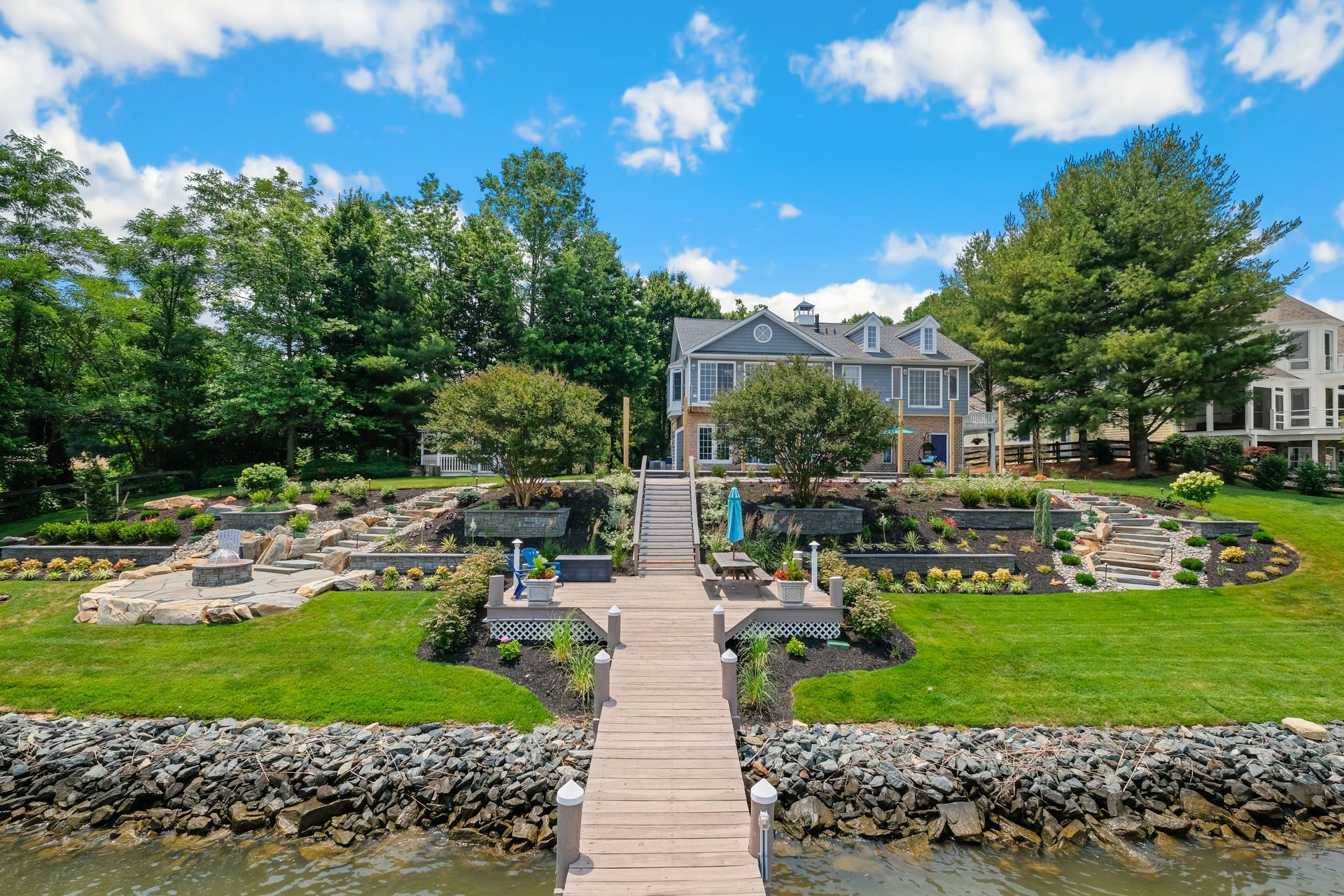 Lakefront garden with stone steps, tiered flower beds, and a large house under a blue sky
