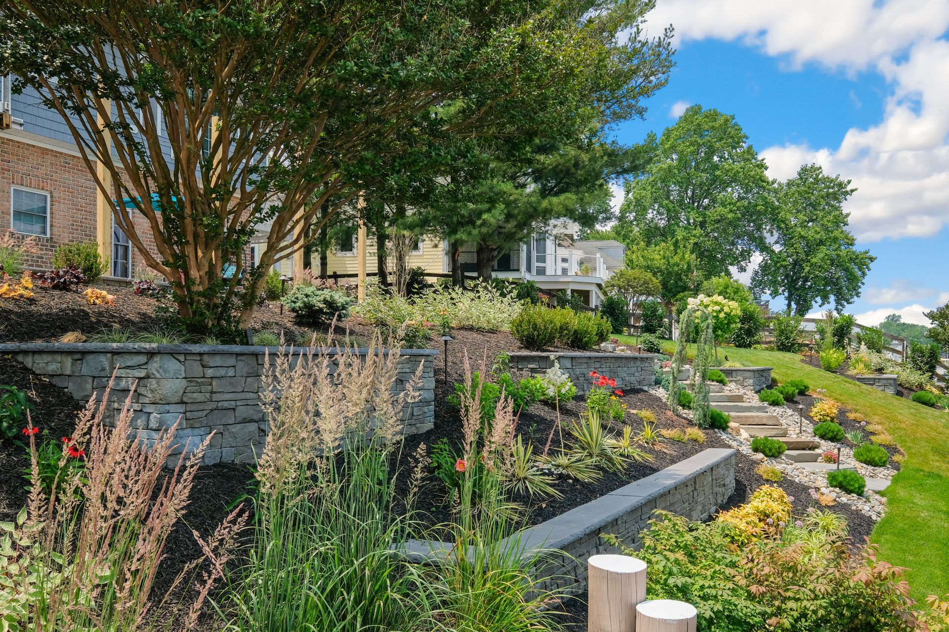 Landscaped garden with stone retaining walls, flowers, and trees beside houses on a sunny day