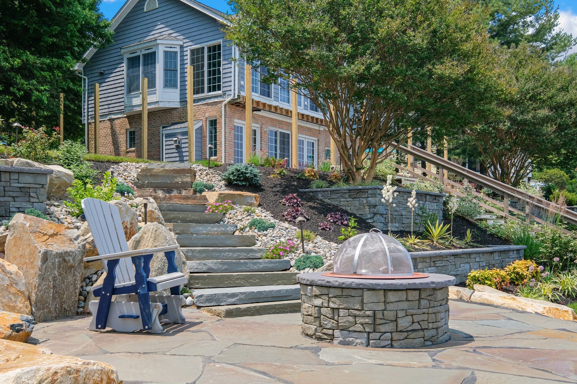 Stone patio with Adirondack chairs, stone steps, and a blue house on a landscaped hillside.