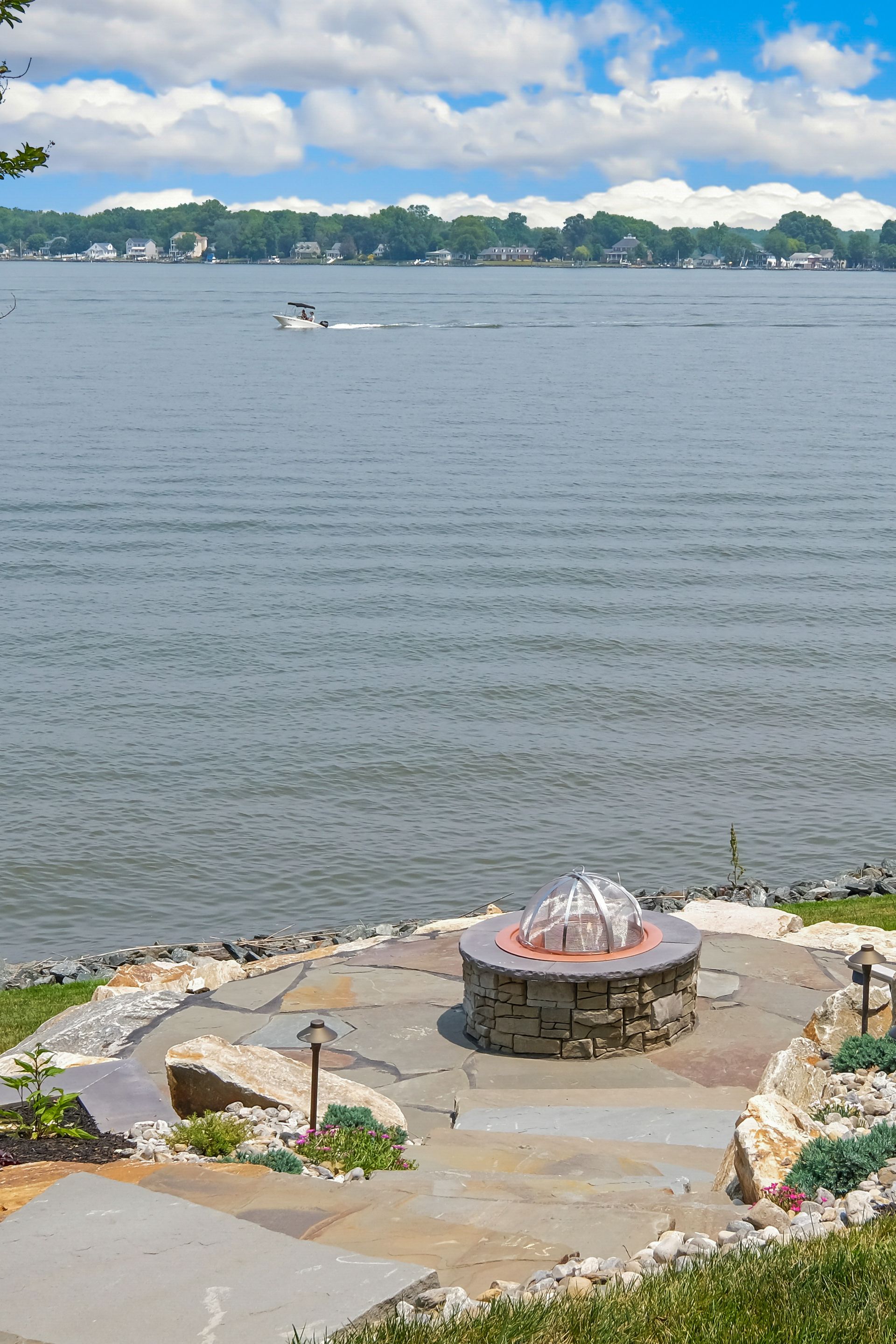 Rocky lakeshore with a small fountain, calm water, and distant houses under a cloudy blue sky