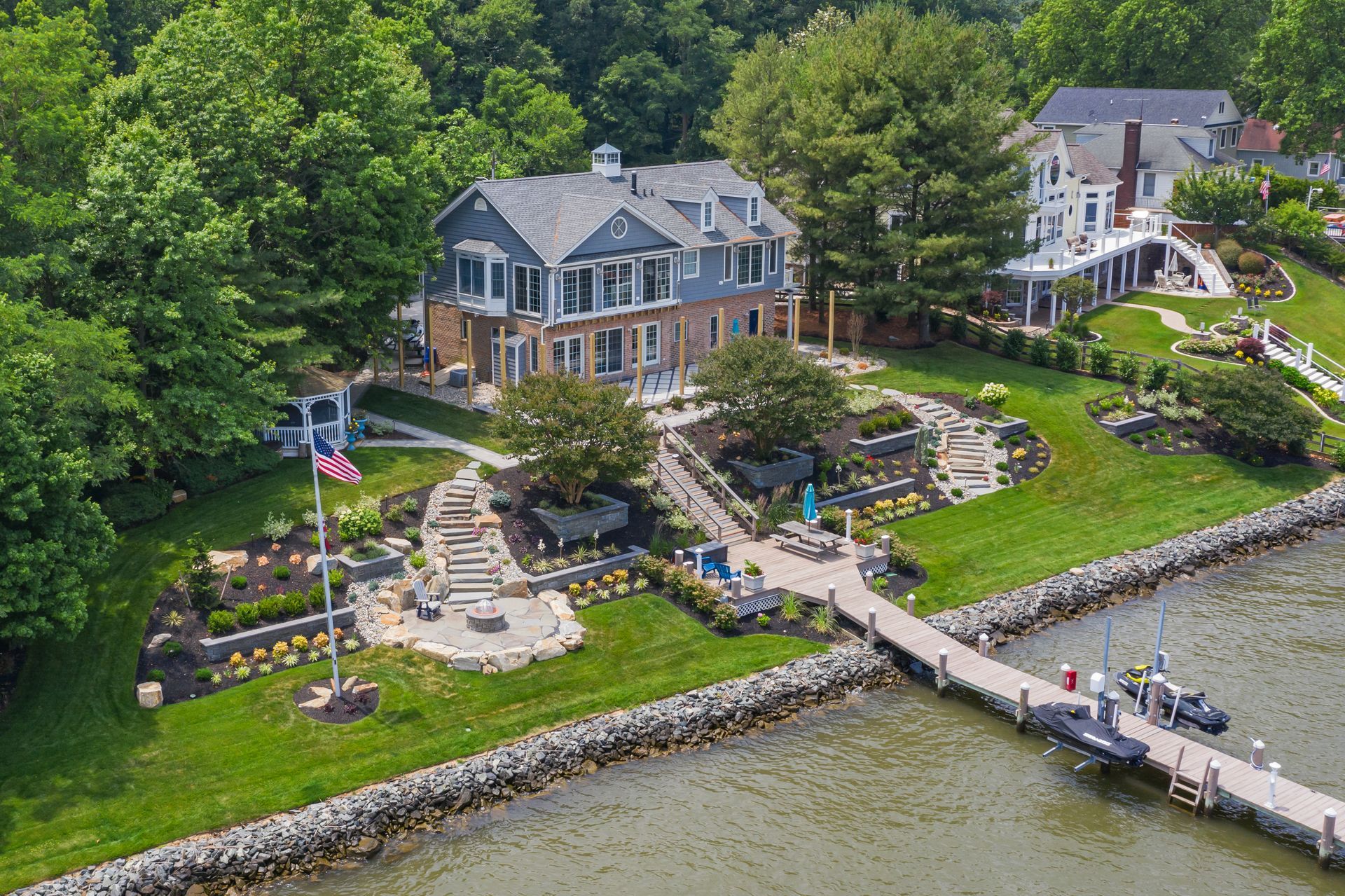 A lakeside stone mansion with gardens, dock, and Canadian flag beside calm water and wooded shoreline