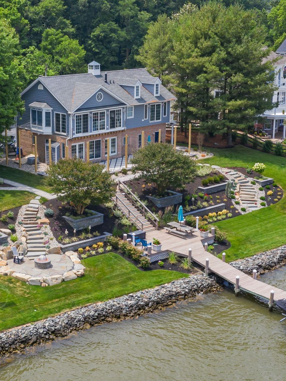 Waterfront stone house with dock, landscaped lawn, and surrounding trees