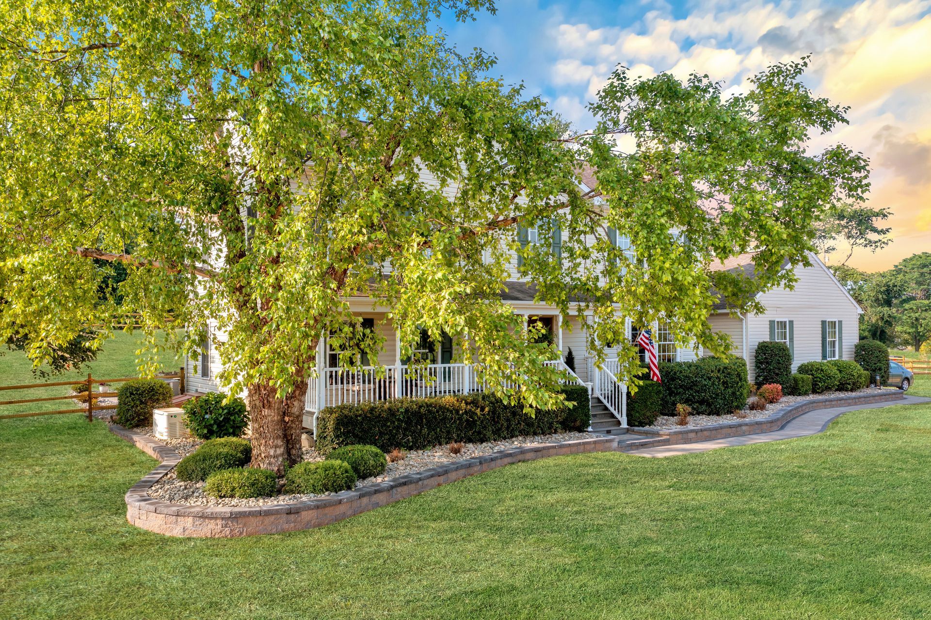 Tree-shaded white house with a wraparound porch, stone edging, and a large green lawn