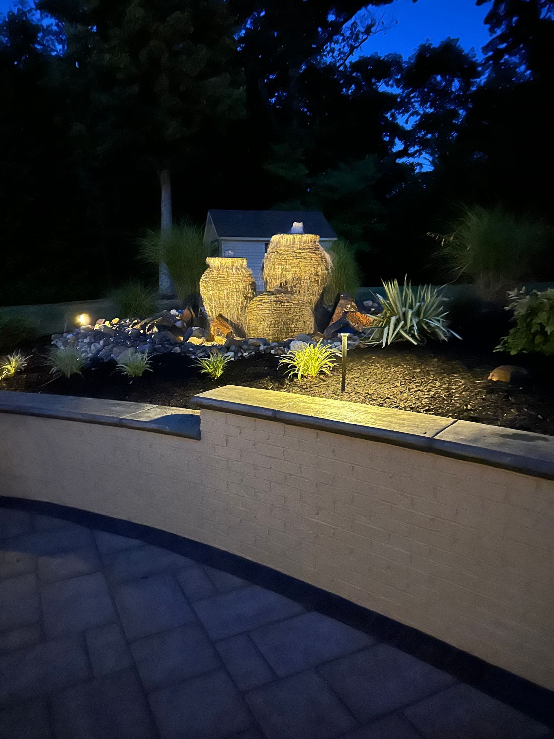 Illuminated garden waterfall and landscaping at dusk behind a curved stone wall