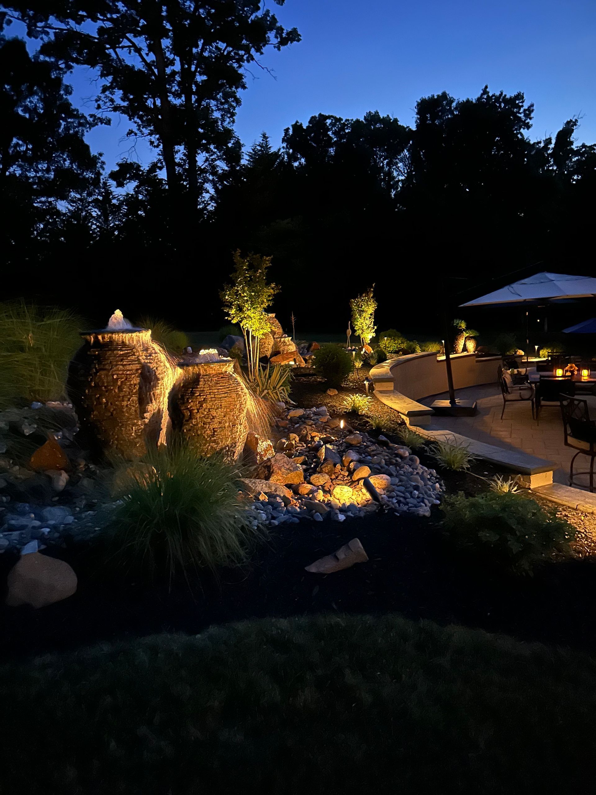 Illuminated backyard waterfall and pond at dusk, with trees silhouetted against the evening sky