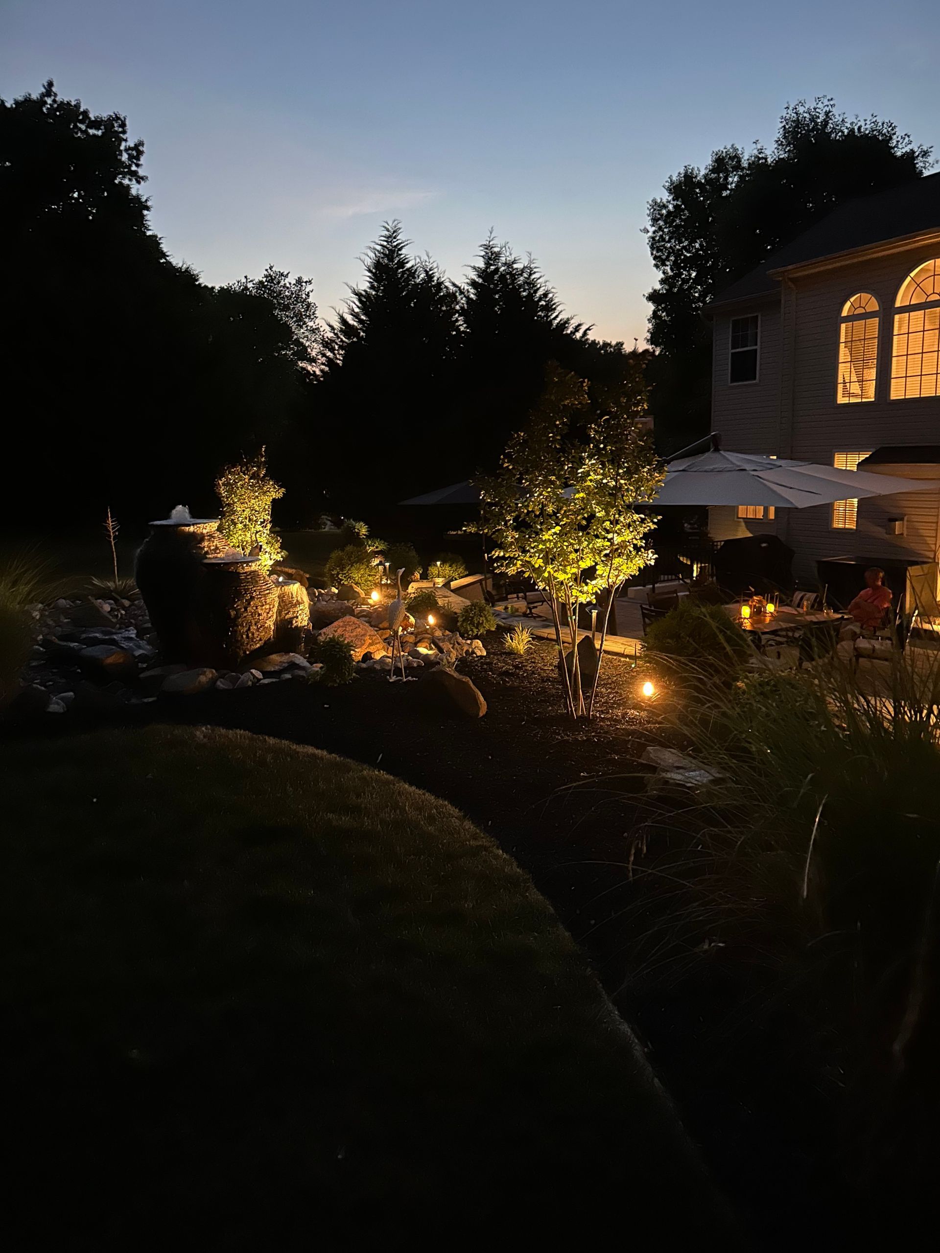 Twilight garden with lit trees, pond, and a house glowing with warm windows and patio lights
