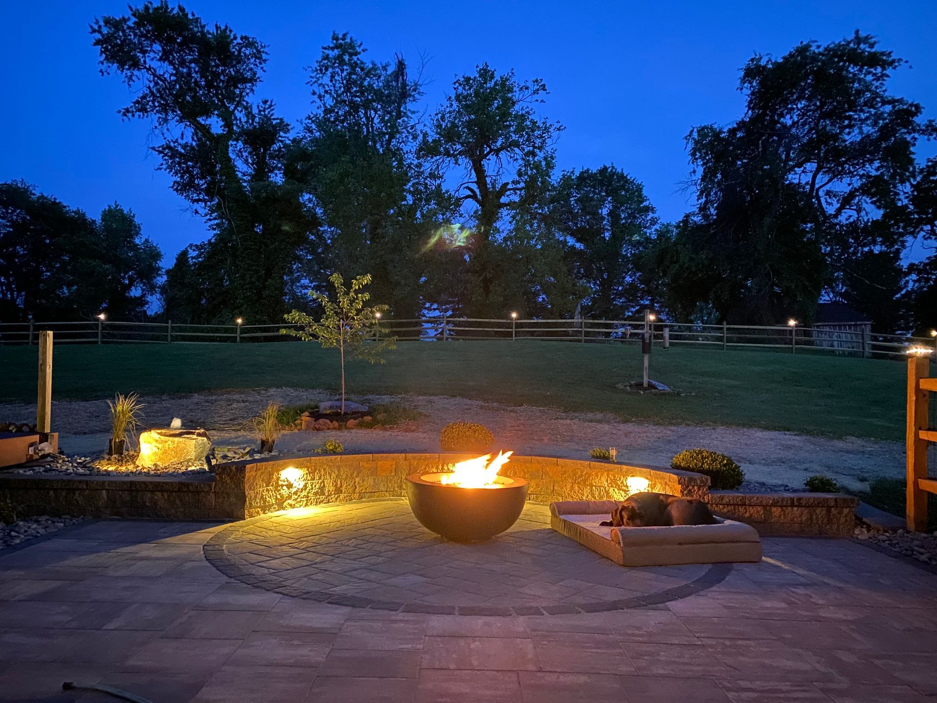 Backyard fire pit area at dusk with glowing flames, stone patio, and trees in the background