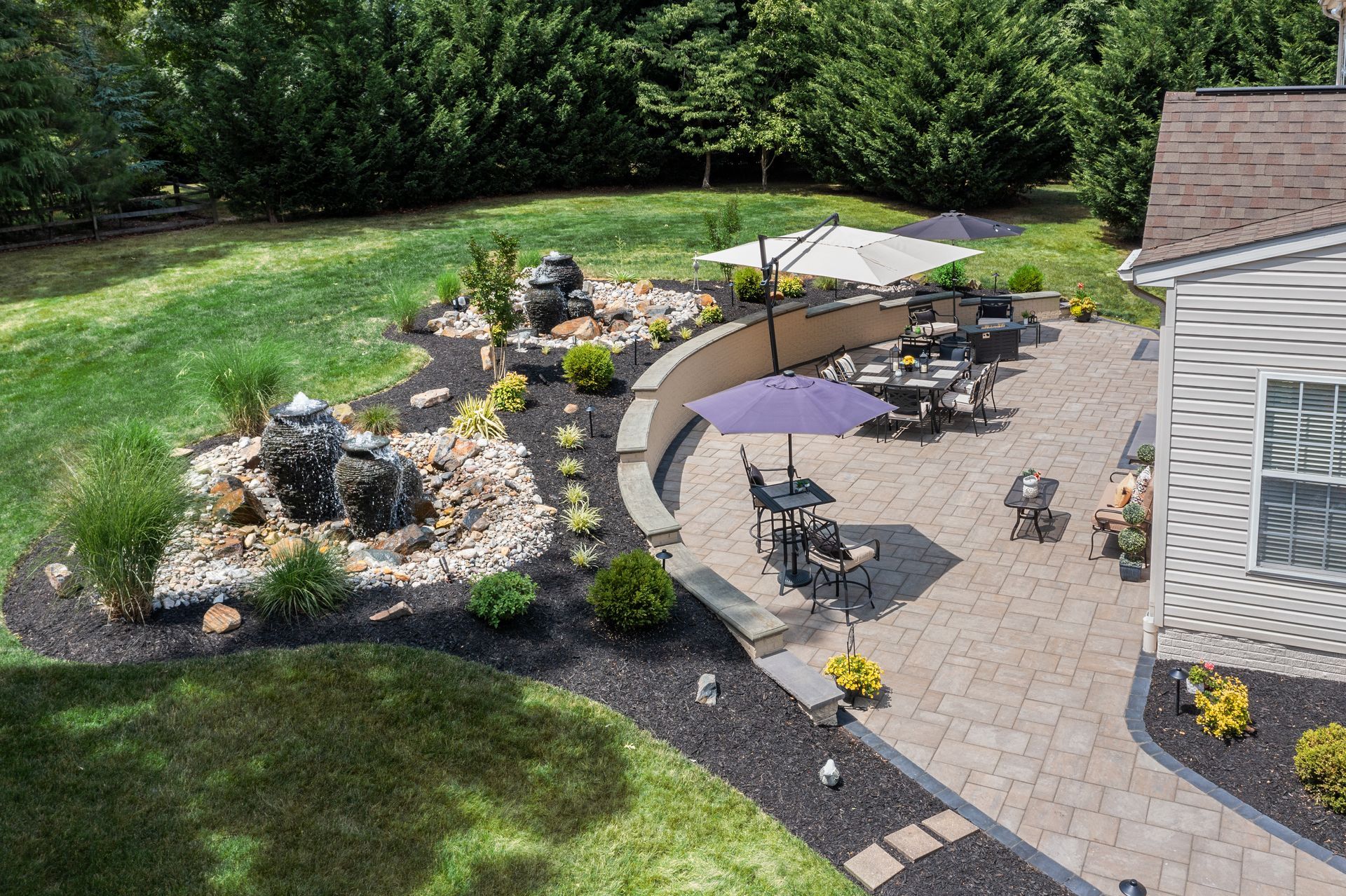 Backyard patio with tables, umbrellas, stone fire pit, and landscaped garden beside a house