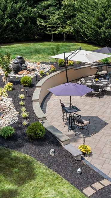 Backyard patio with tables, umbrellas, stone fire pit, and landscaped garden beside a house