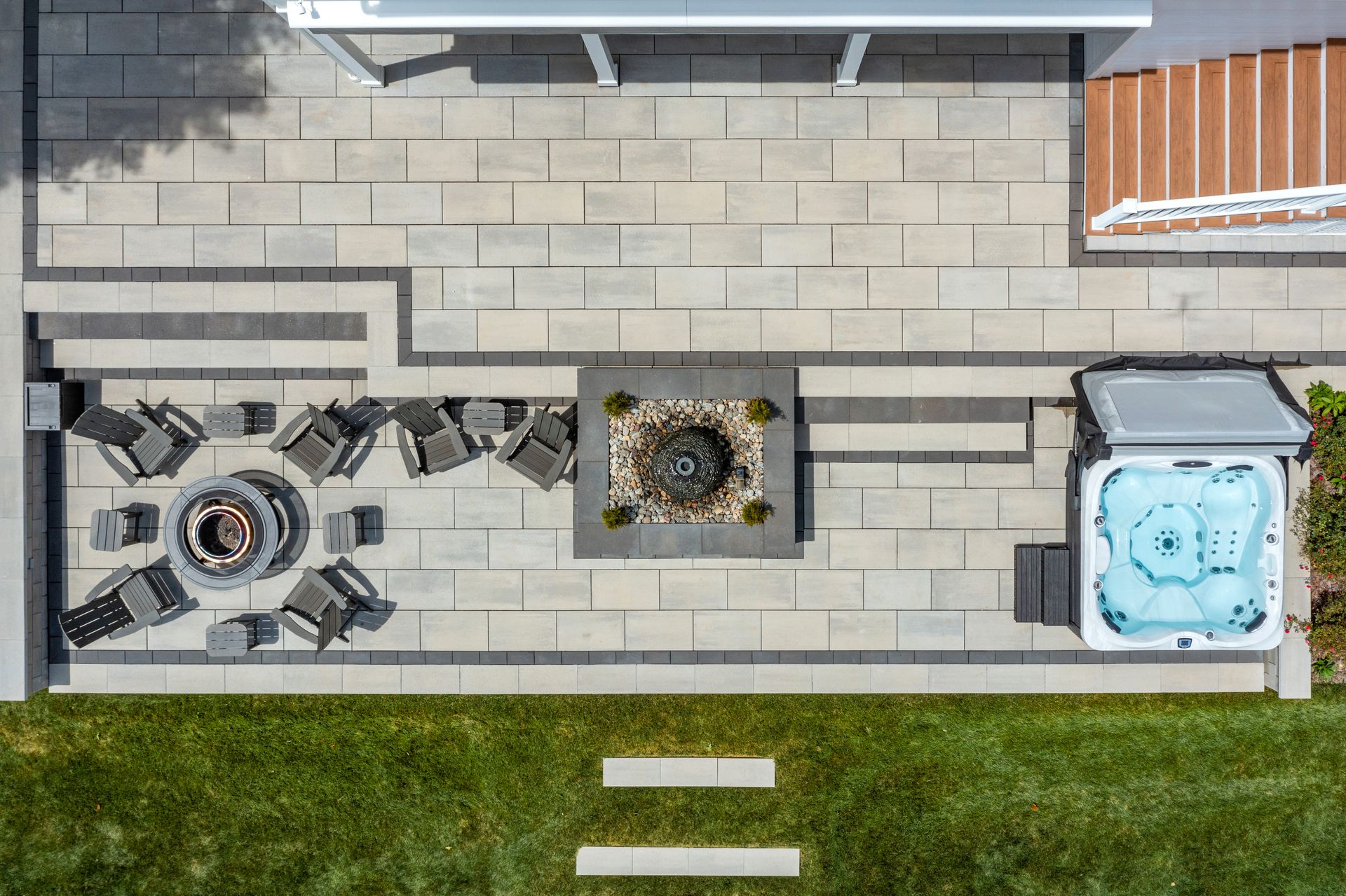 Overhead view of a backyard patio with stone paving, fire pit seating, and a small hot tub beside a grass lawn