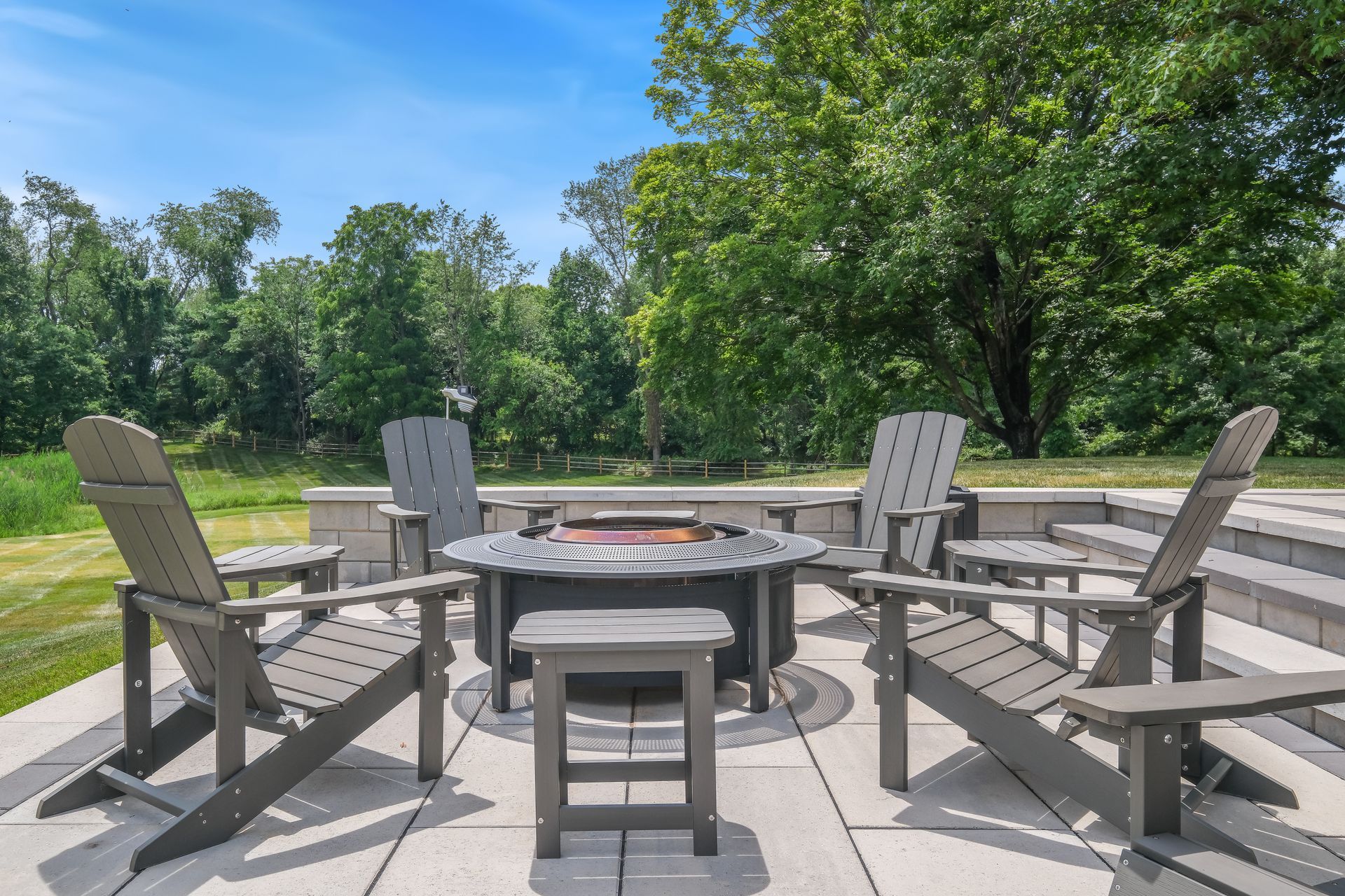 Outdoor fire pit seating area with Adirondack chairs on a patio, surrounded by trees and grass.