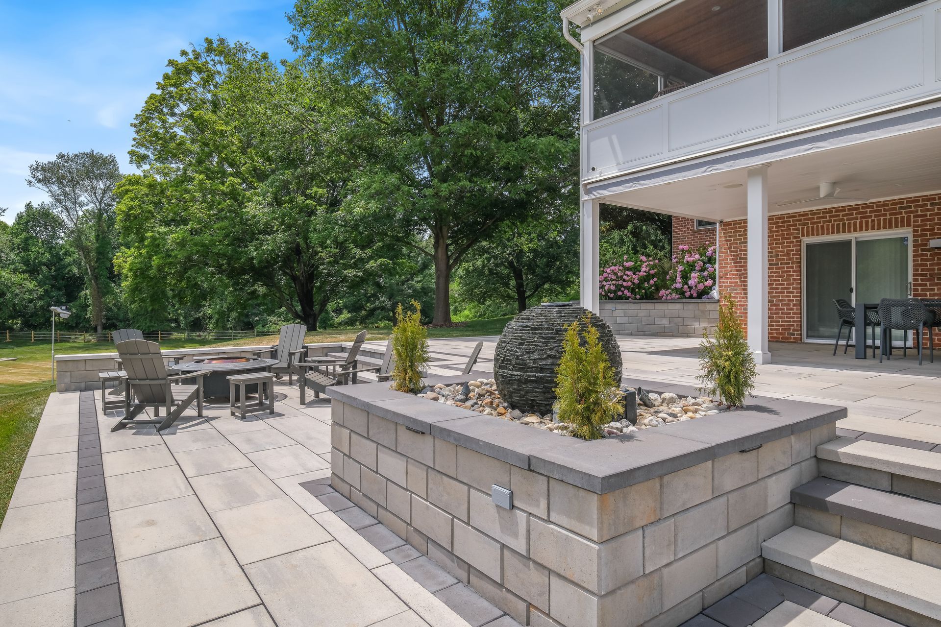 Patio with stone seating, tables, steps, and a covered porch beside a grassy yard and trees.