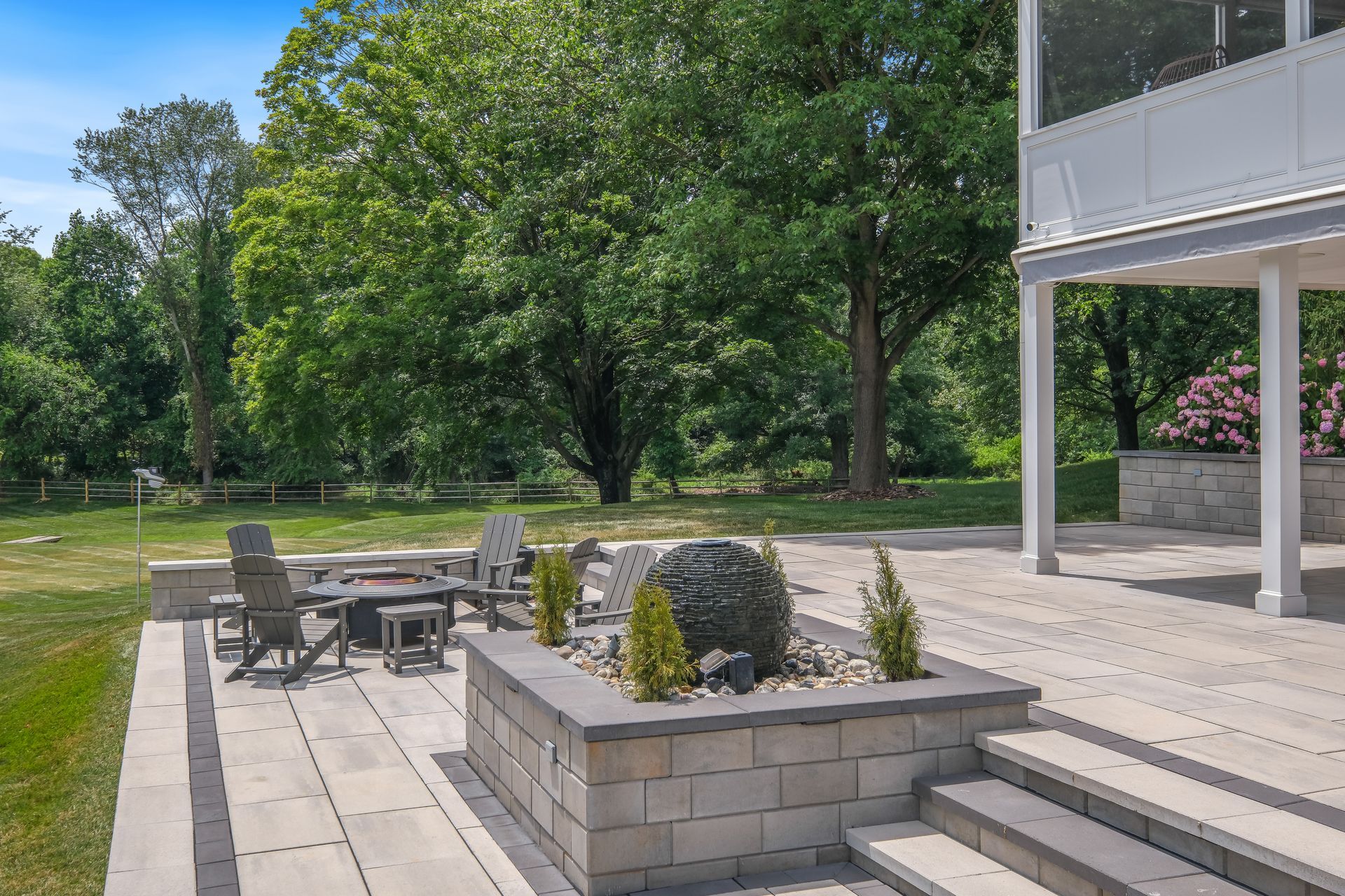 Patio with stone planters, outdoor seating, and steps beside a white house, bordered by green trees