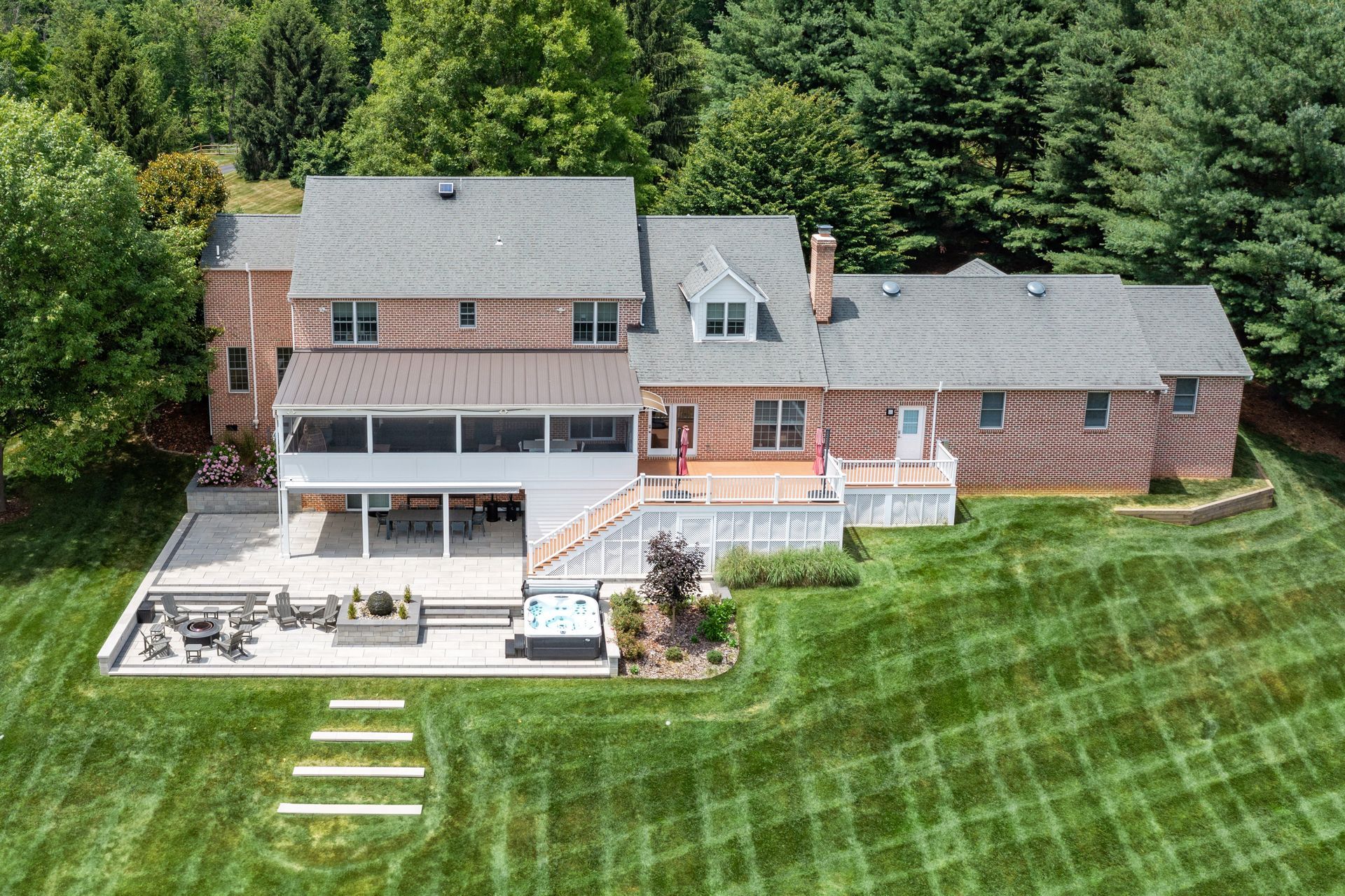 Aerial view of a large brick house with a deck and patio on a green lawn, backed by dense trees