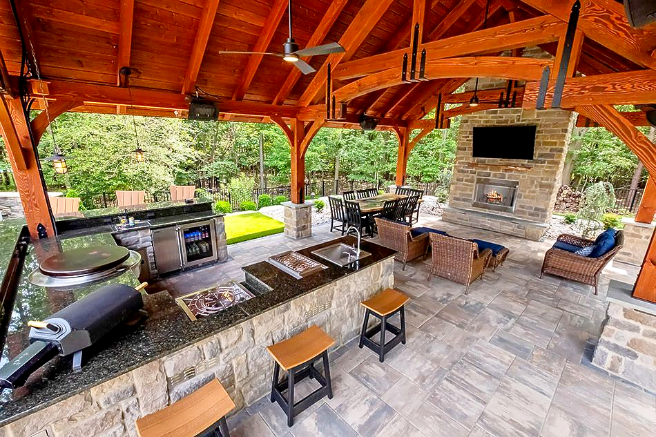 Covered outdoor kitchen and patio with stone fireplace, grill, and seating under a wooden roof