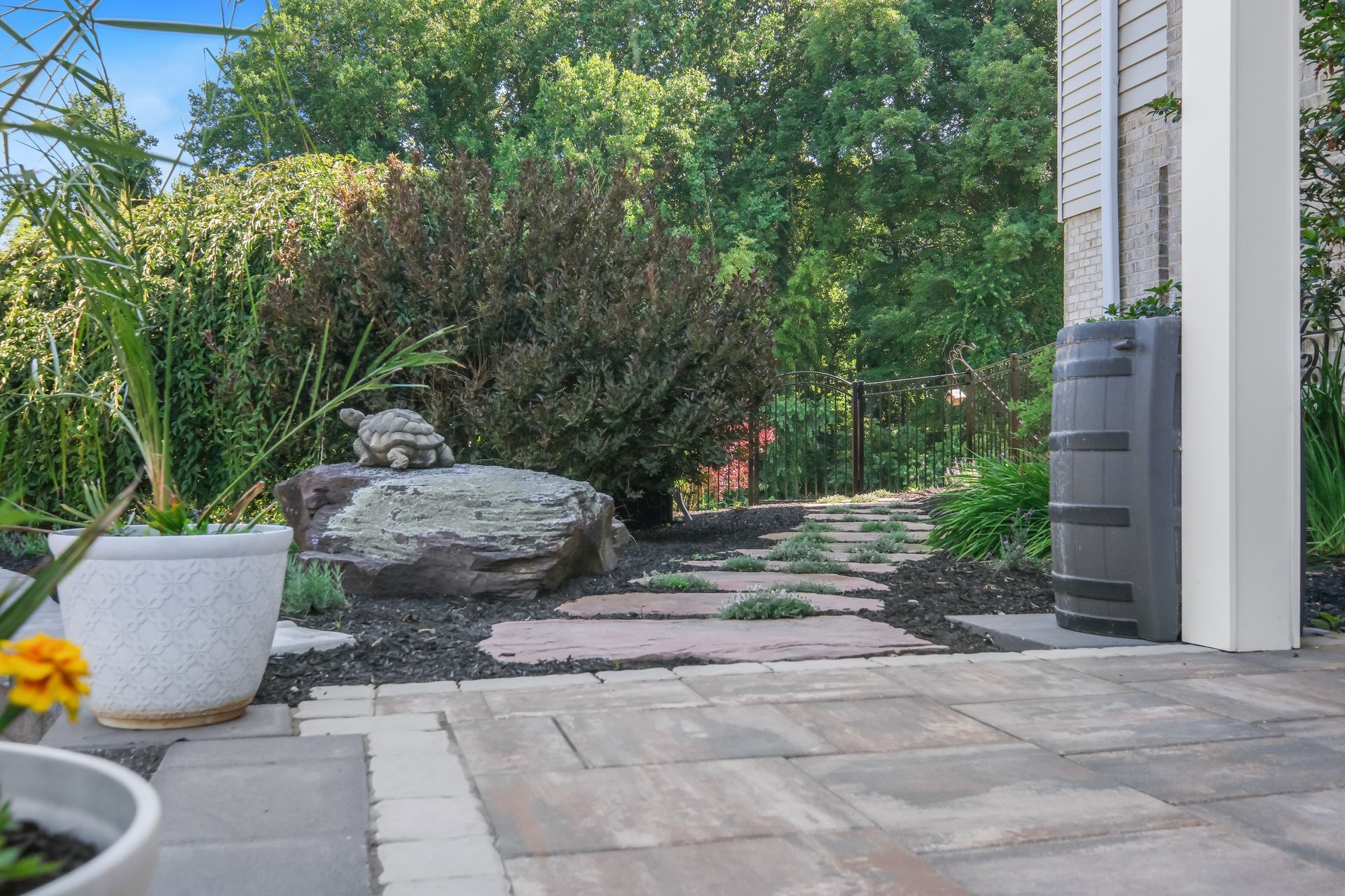 Stone garden path beside patio with potted plants and a large rock, leading into lush greenery