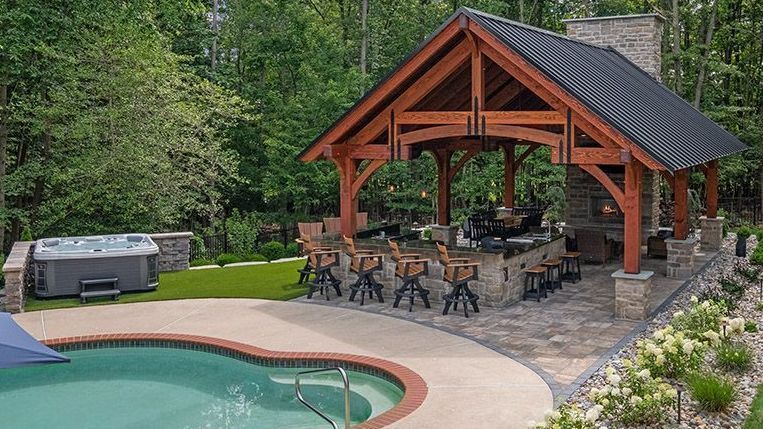 Backyard patio with a wooden pavilion, outdoor dining set, hot tub, and swimming pool beside trees.
