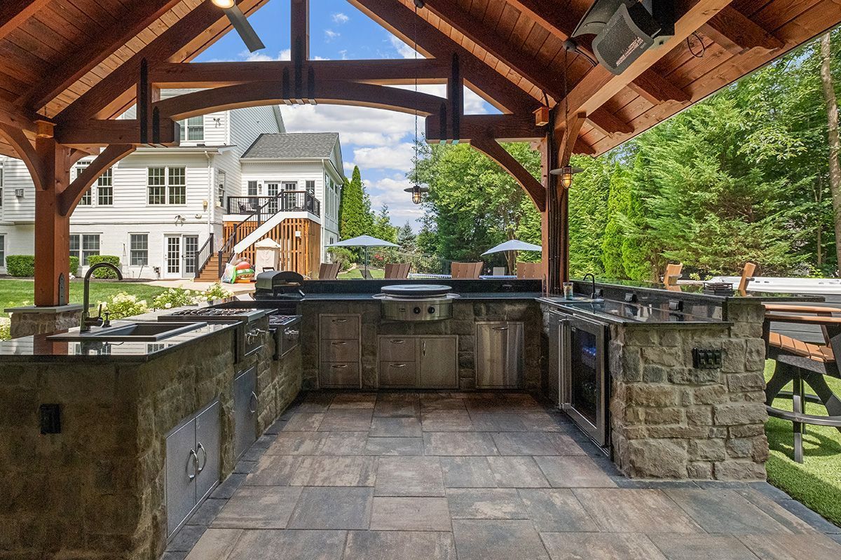 Covered outdoor stone kitchen and grill area with wooden roof, overlooking a house and trees