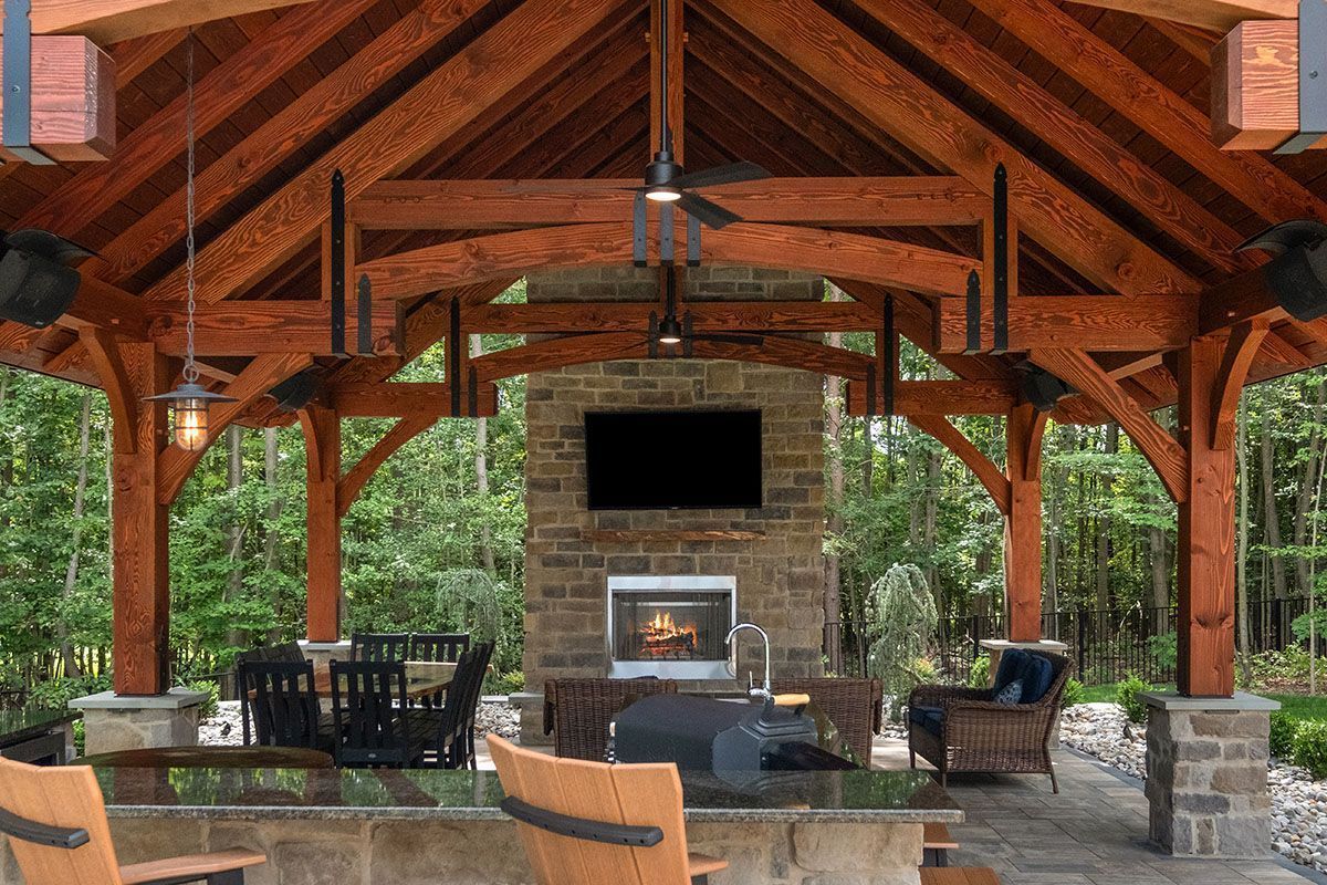 Rustic outdoor pavilion with stone fireplace, wooden beams, and seating on a patio surrounded by greenery